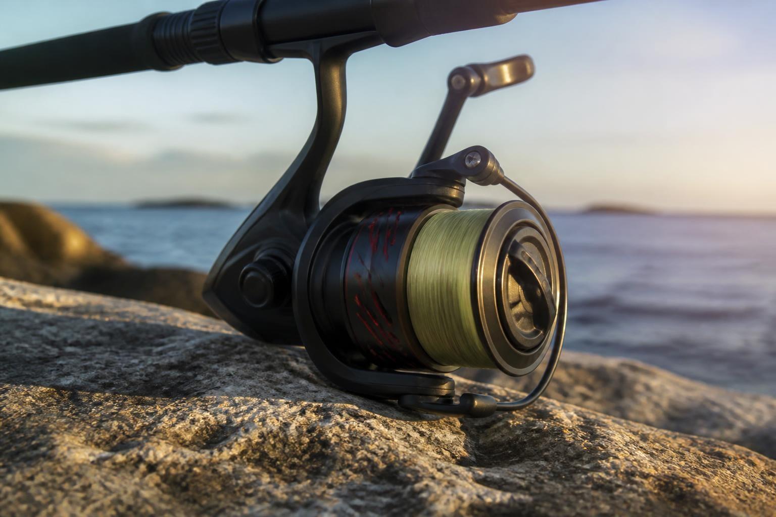 Spinning reel spooled with green braided line, mounted on a rod resting on shoreline rocks by the sea