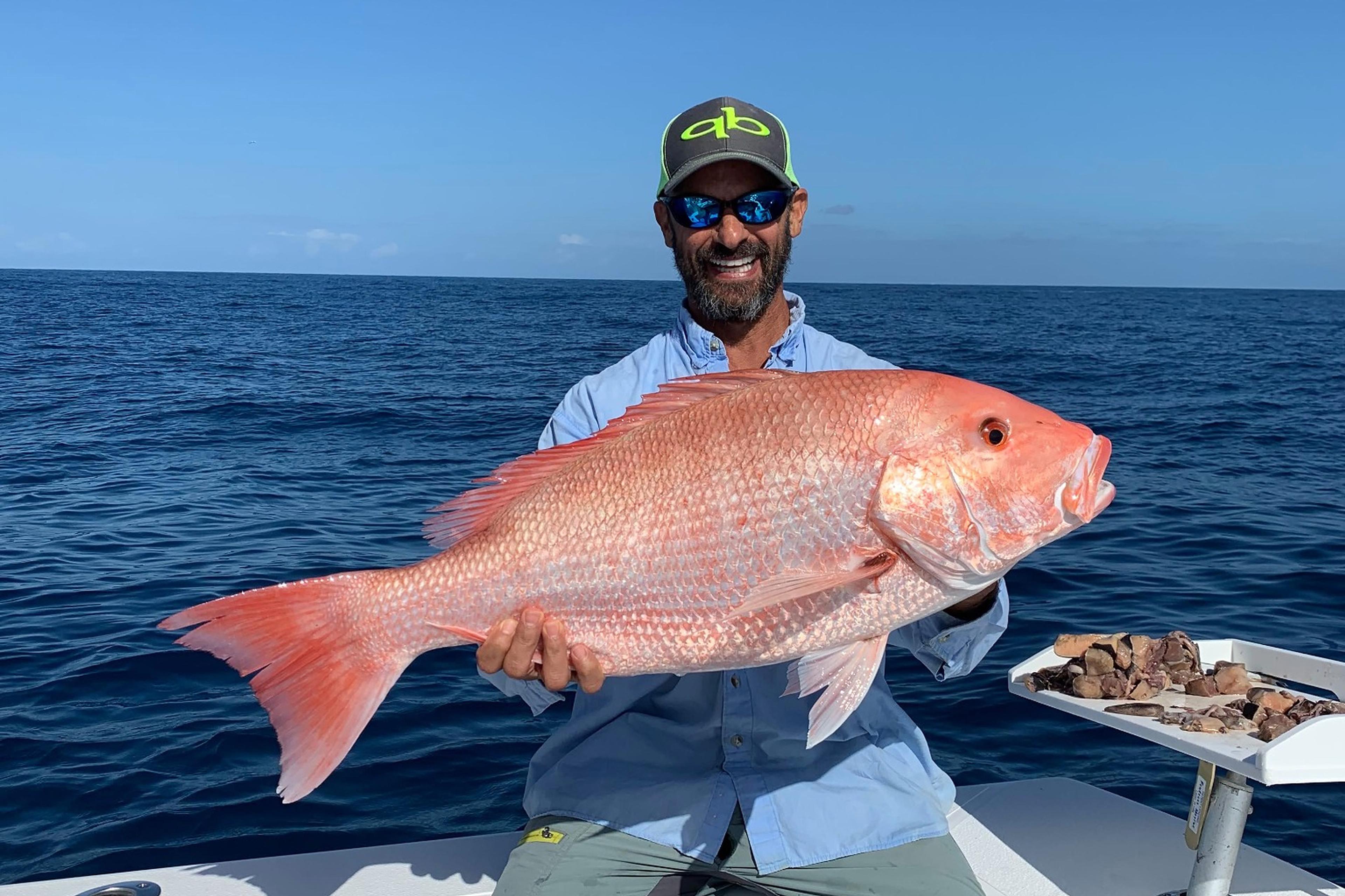 A smiling angler in a cap and sunglasses holds up a large red snapper on a boat offshore, with deep blue ocean and clear sky behind.