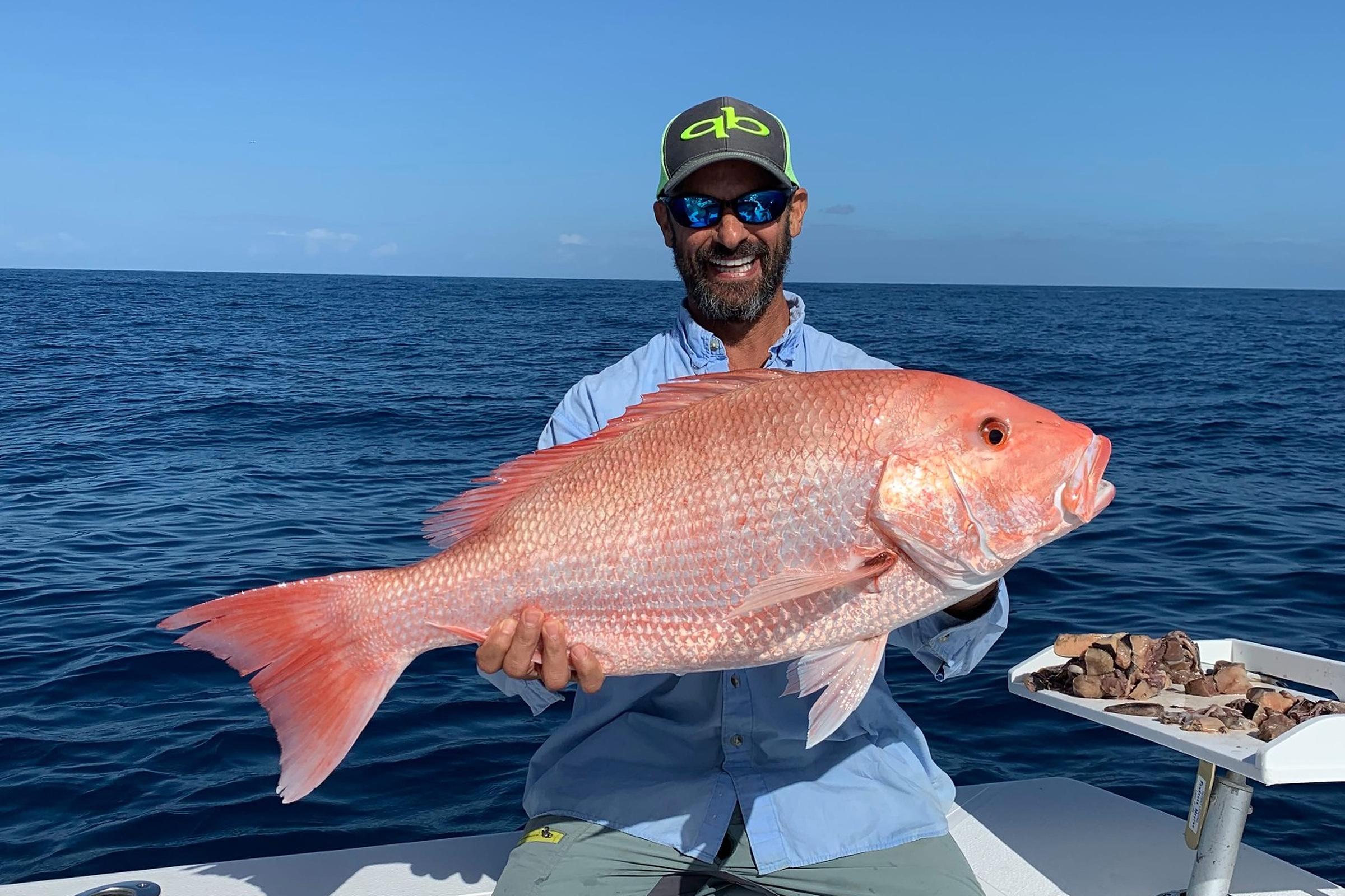 A smiling angler in a cap and sunglasses holds up a large red snapper on a boat offshore, with deep blue ocean and clear sky behind.