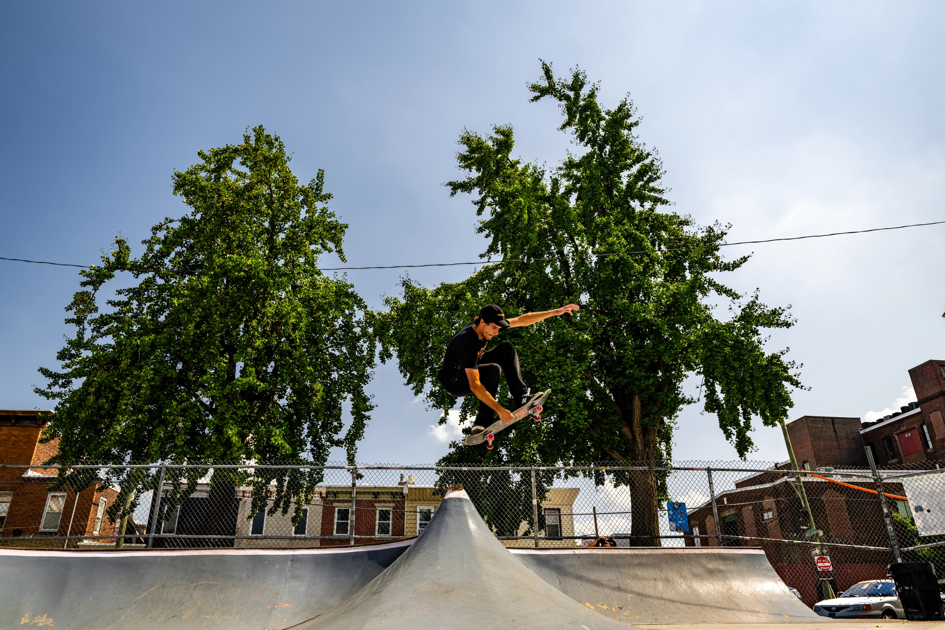 A skateboarder performs a trick mid-air above a concrete ramp, with large green trees and brick buildings framing the outdoor skate park.