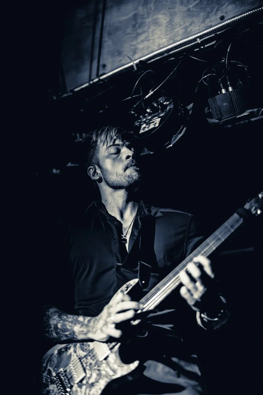 A tattooed guitarist performs on stage in low blue light, eyes closed, hands on bass guitar with overhead stage rigging visible.