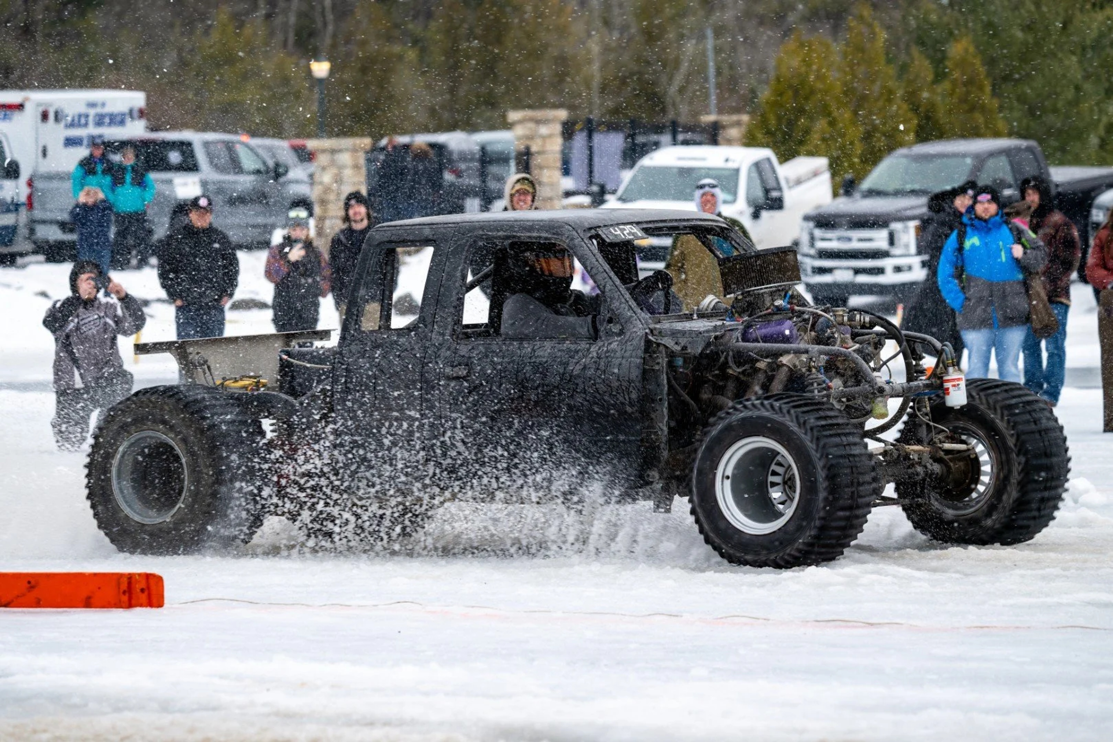 A custom hot rod with exposed engine accelerates on snow, kicking up a white cloud, with spectators watching from the sidelines at a winter event.