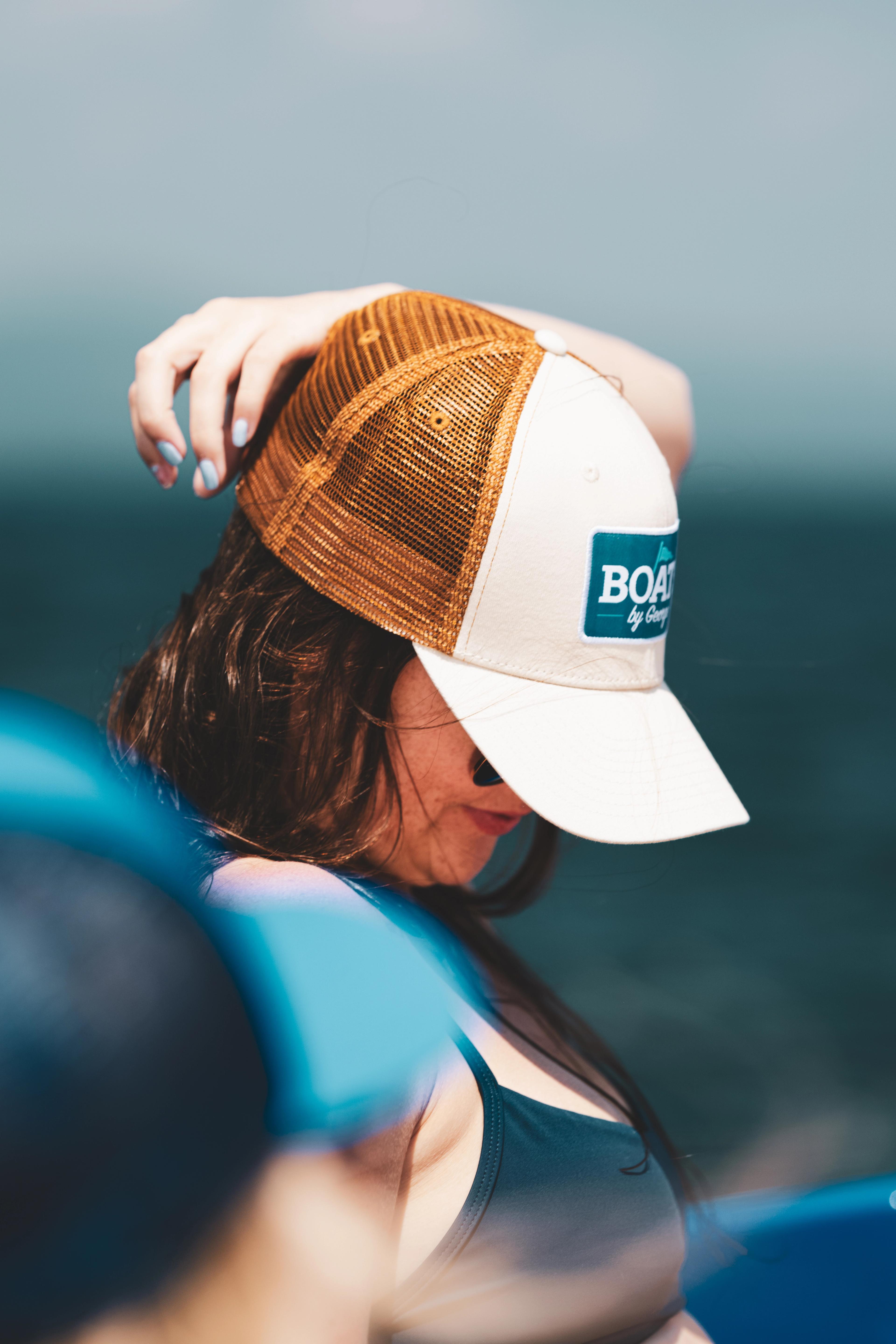 A person wearing a white and tan baseball cap gazes toward open water, shot from behind with a blurred blue kayak in the foreground.