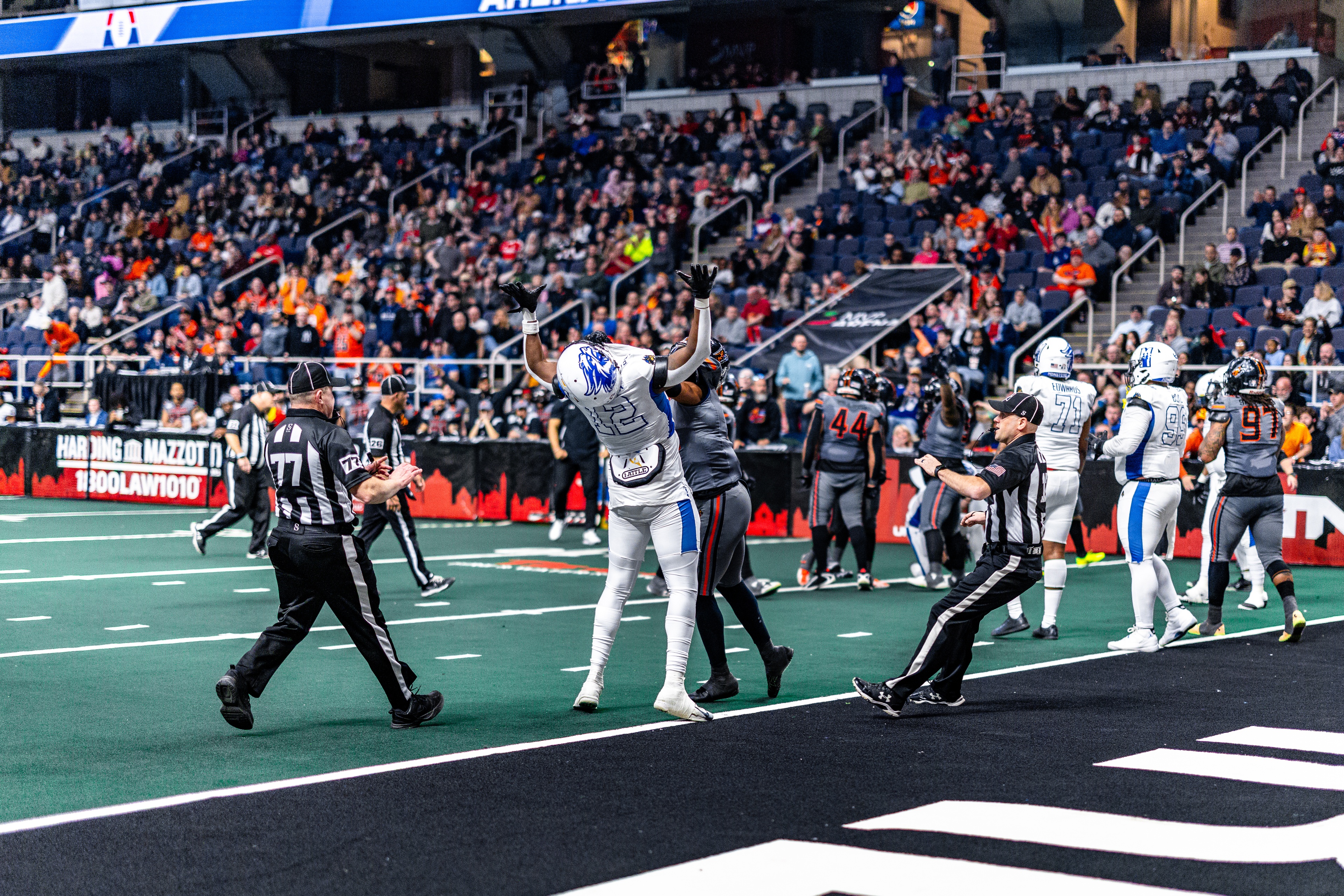 An arena football player in white and blue uniform celebrates with arms raised on the field, surrounded by opposing players and officials with a packed stadium crowd in the background.