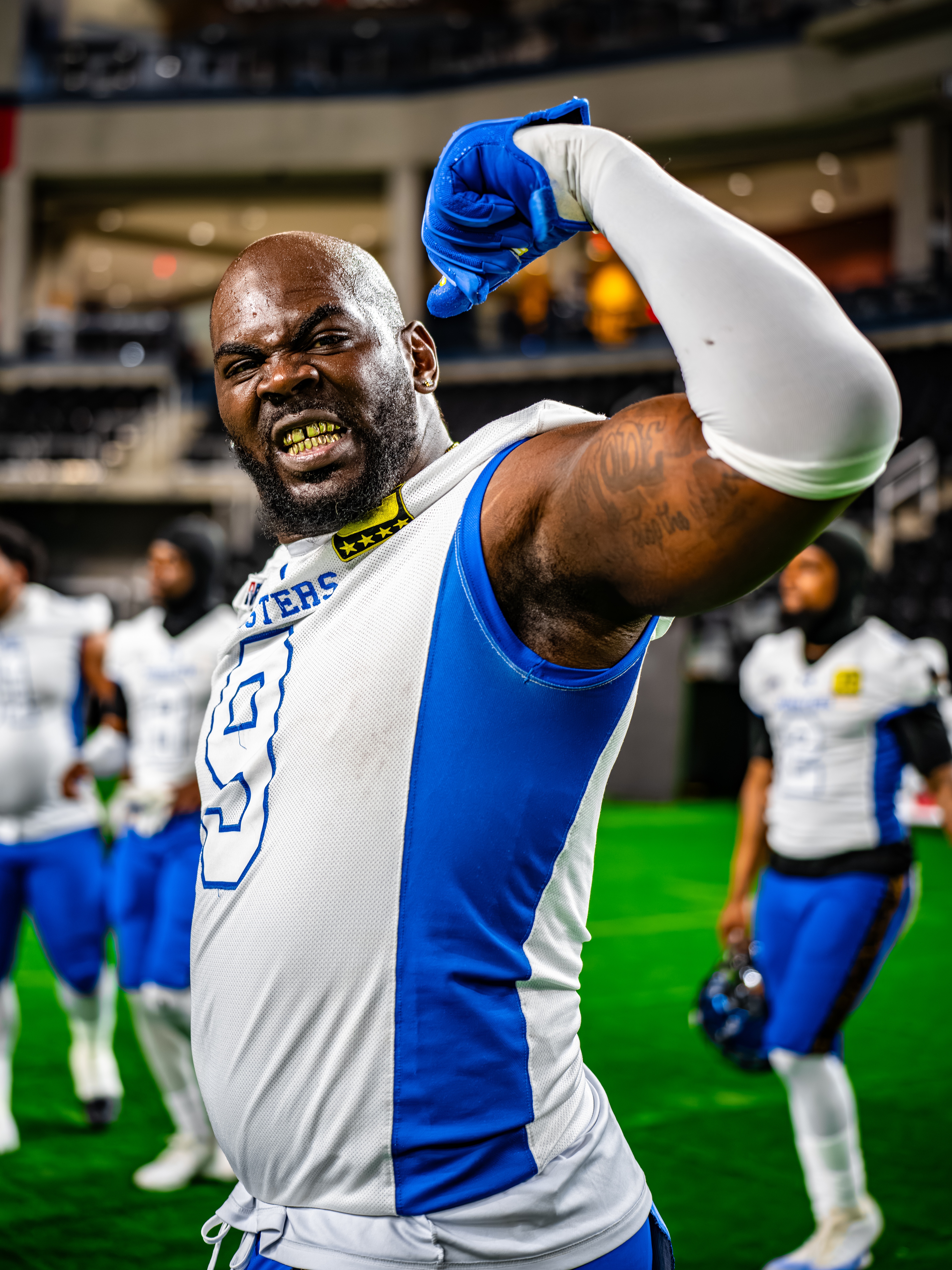 A muscular athlete in blue and white uniform raises a blue-gloved fist in celebration on an indoor football field, smiling with intensity.