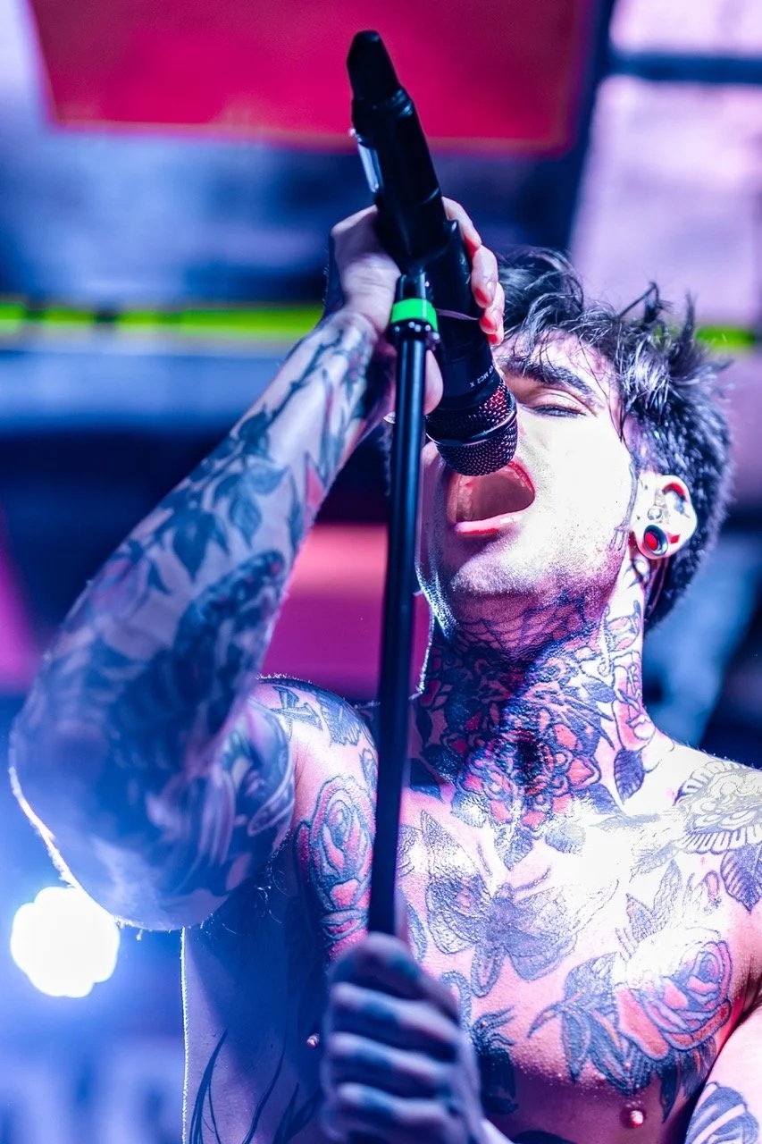 A tattooed singer on stage grips a microphone with intensity, bathed in pink and blue stage lighting, eyes closed in performance.