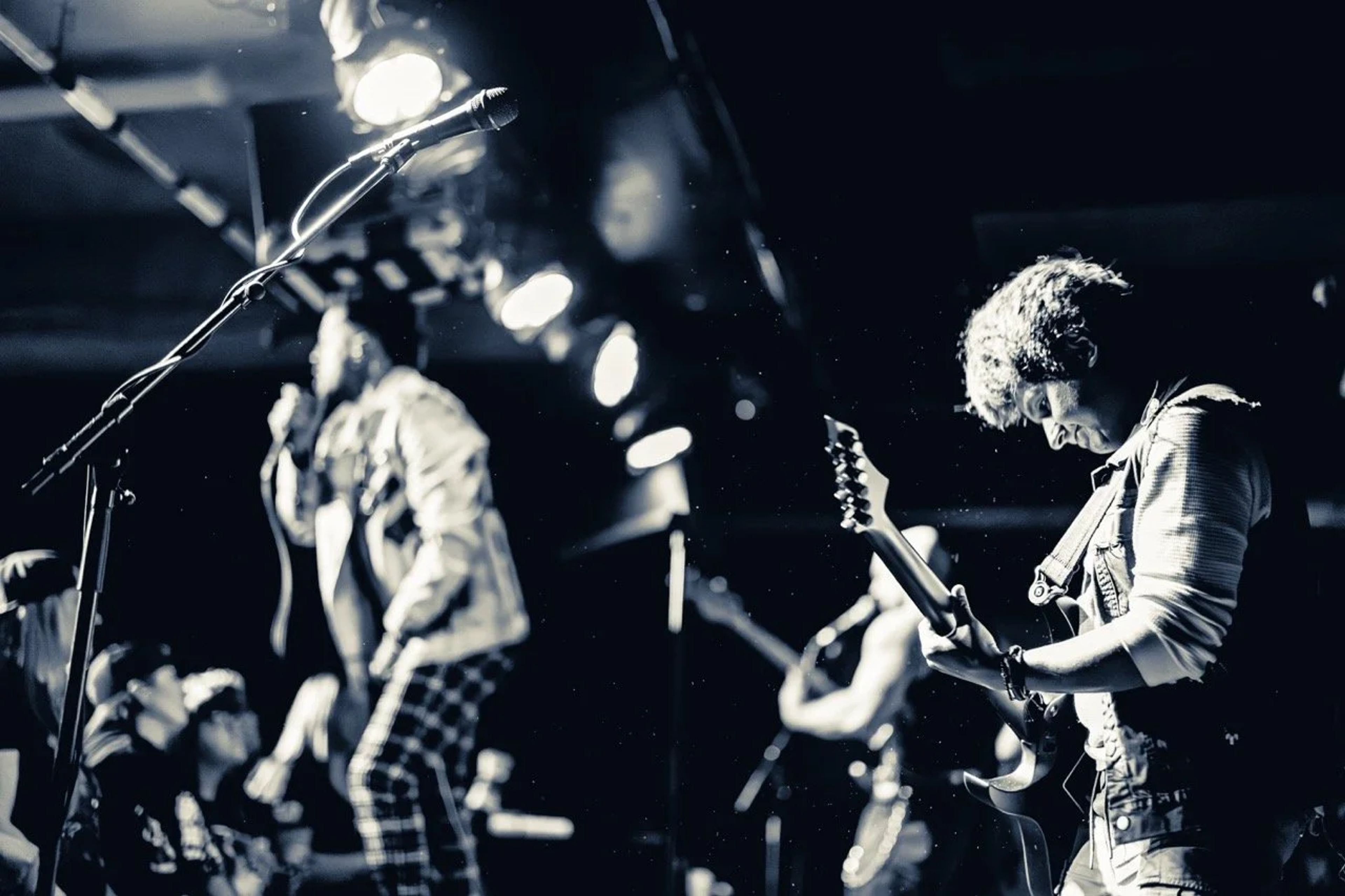 Two guitarists performing on stage at Empire Live Underground, lit in stark white and black contrast, with stage lights and microphone visible in the foreground.