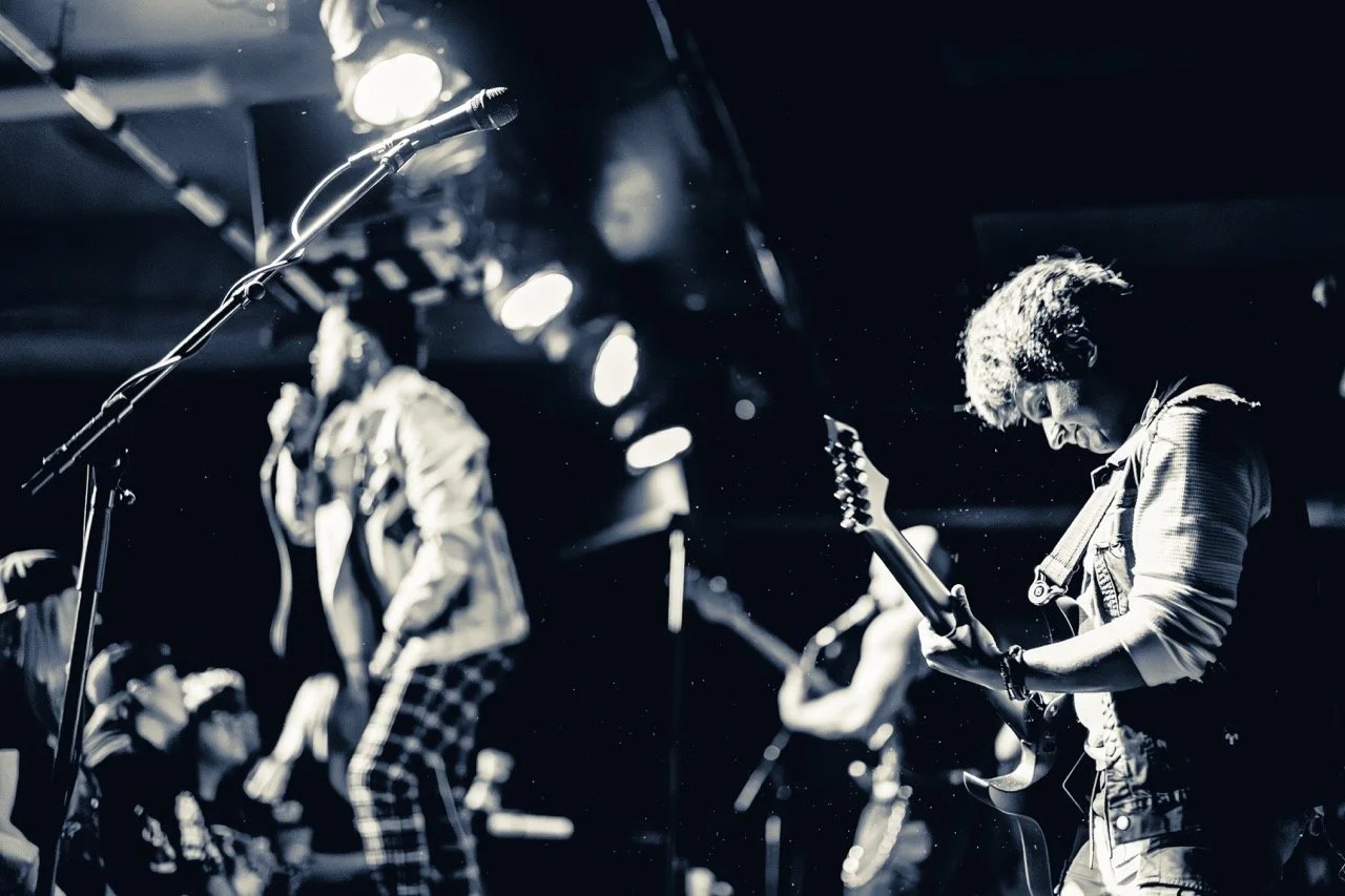 Two guitarists performing on stage at Empire Live Underground, lit in stark white and black contrast, with stage lights and microphone visible in the foreground.