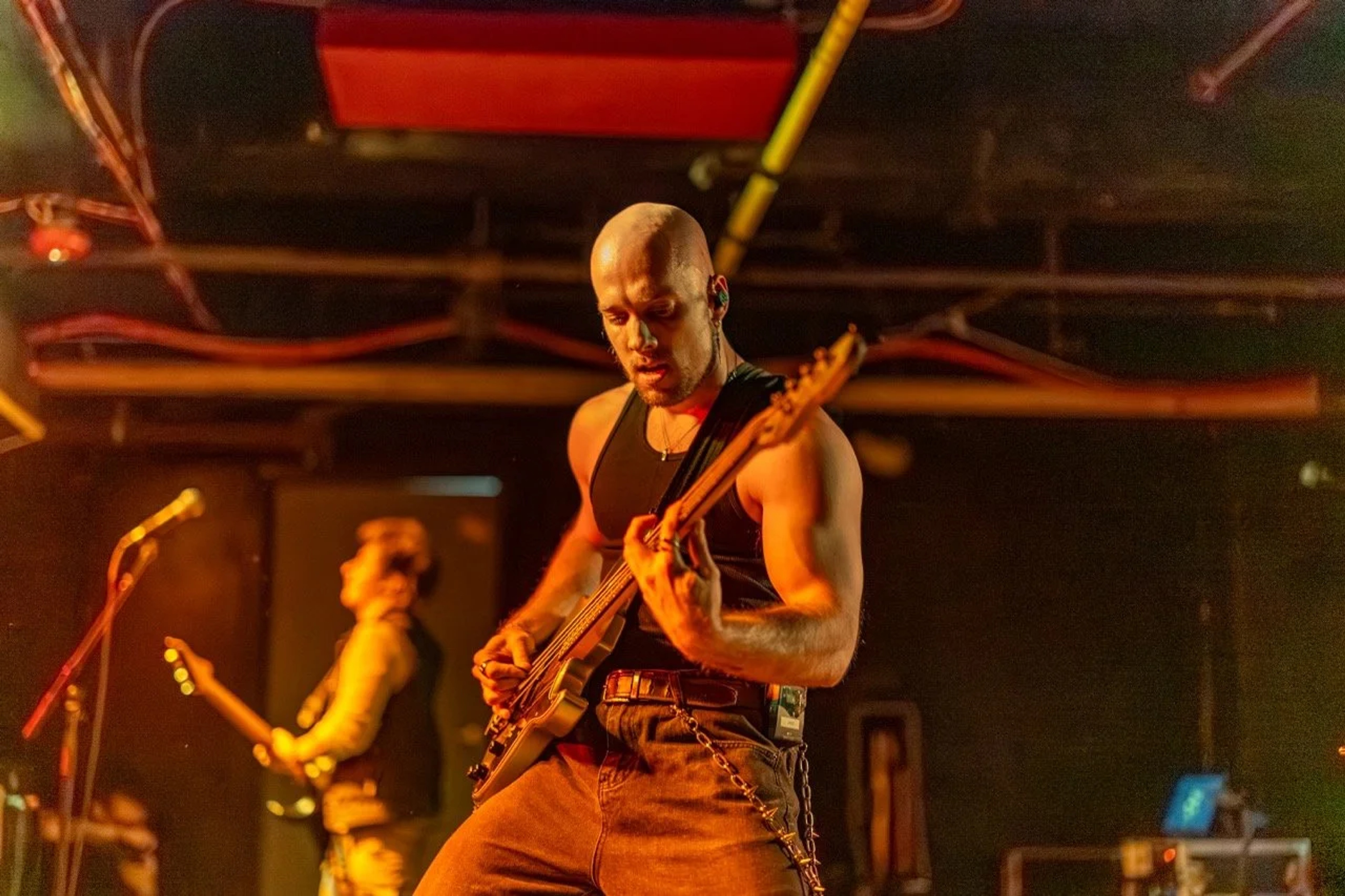 A shirtless bassist performs on stage under warm orange and red lighting, focused on his instrument with a drummer visible in the background.
