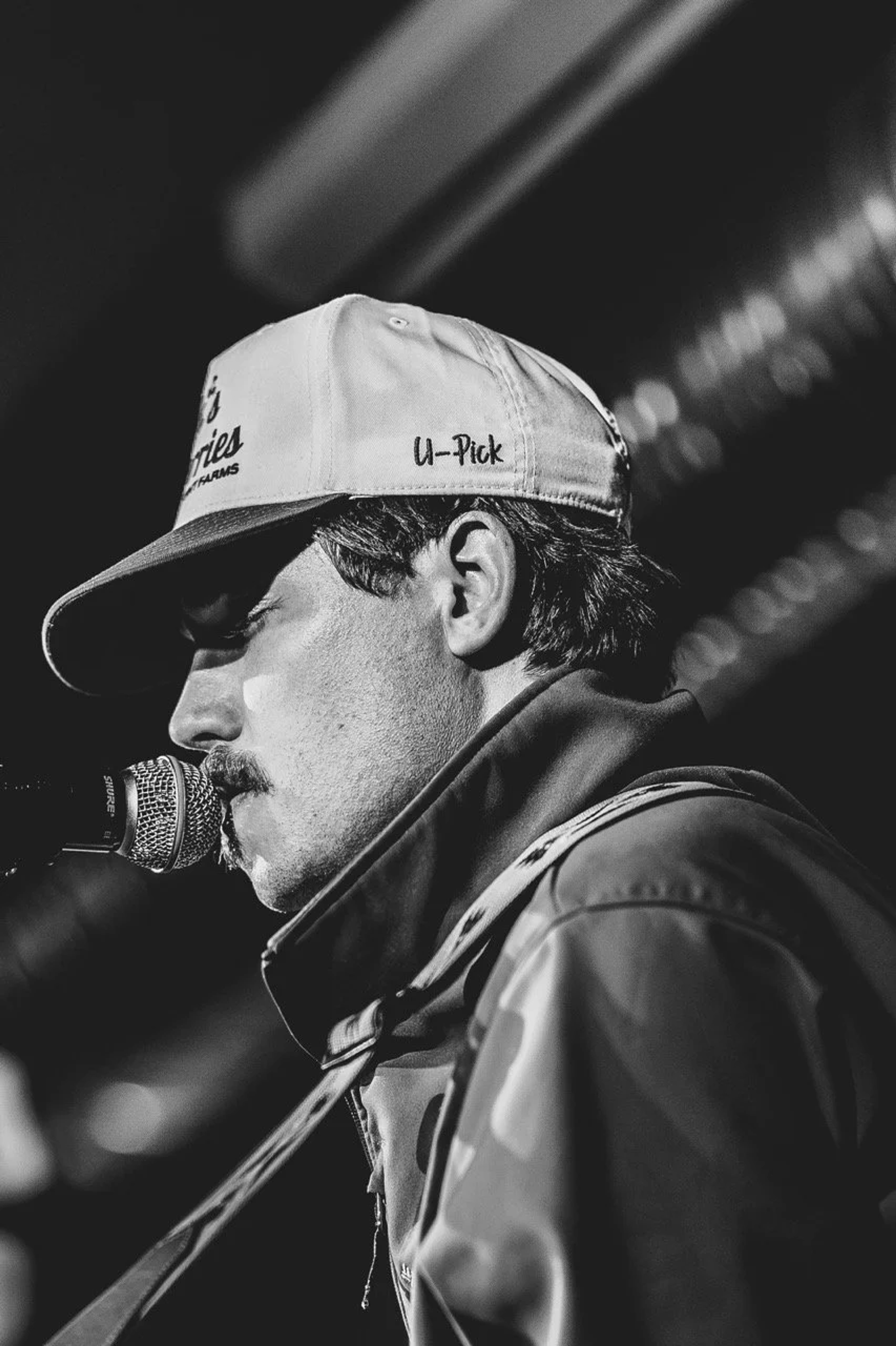 A musician in profile wearing a U-Pick cap, holding a microphone, captured in black and white under stage lighting at an indoor venue.