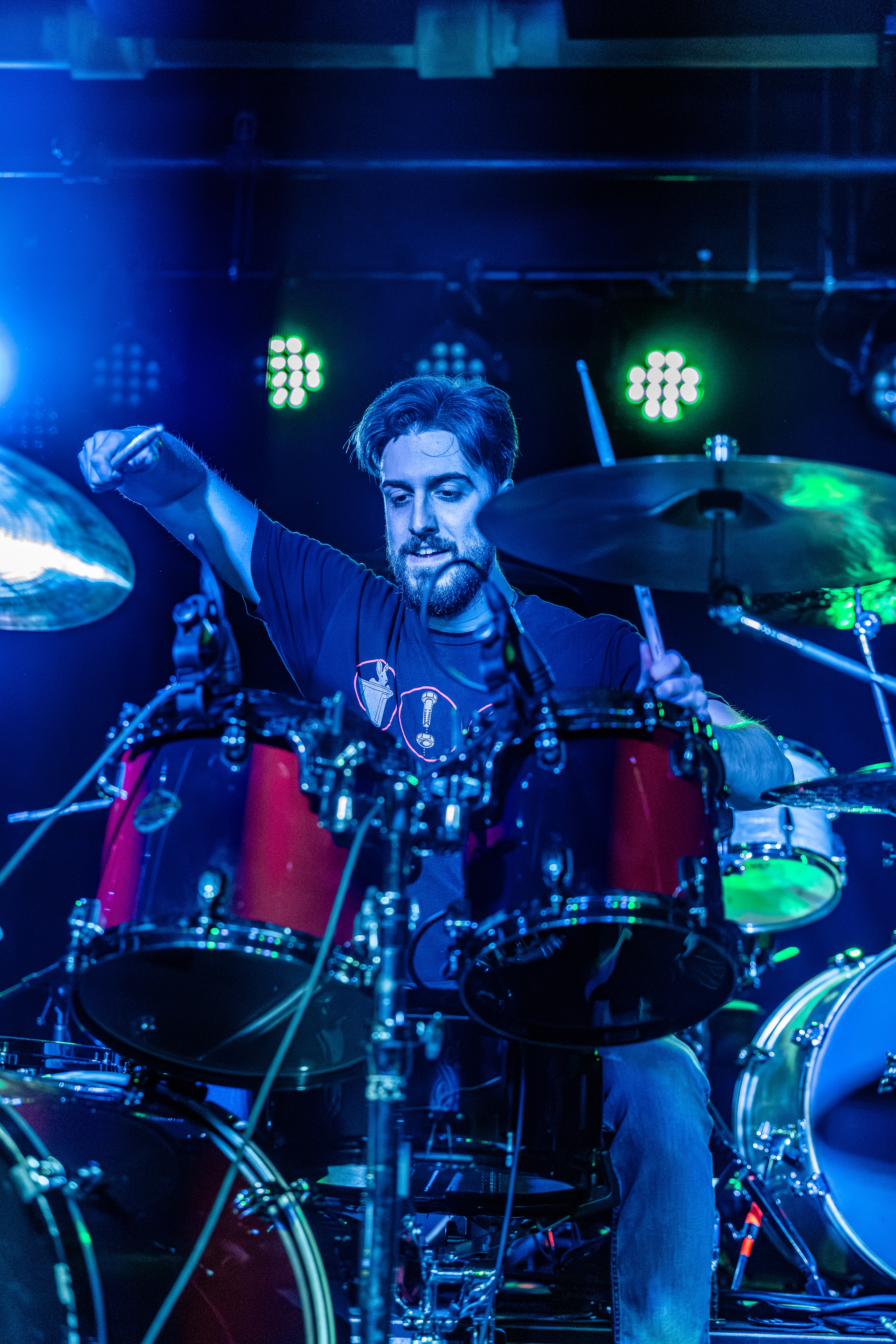 A drummer on stage behind red drums, fist raised, bathed in blue and green stage lighting with drum kit visible.