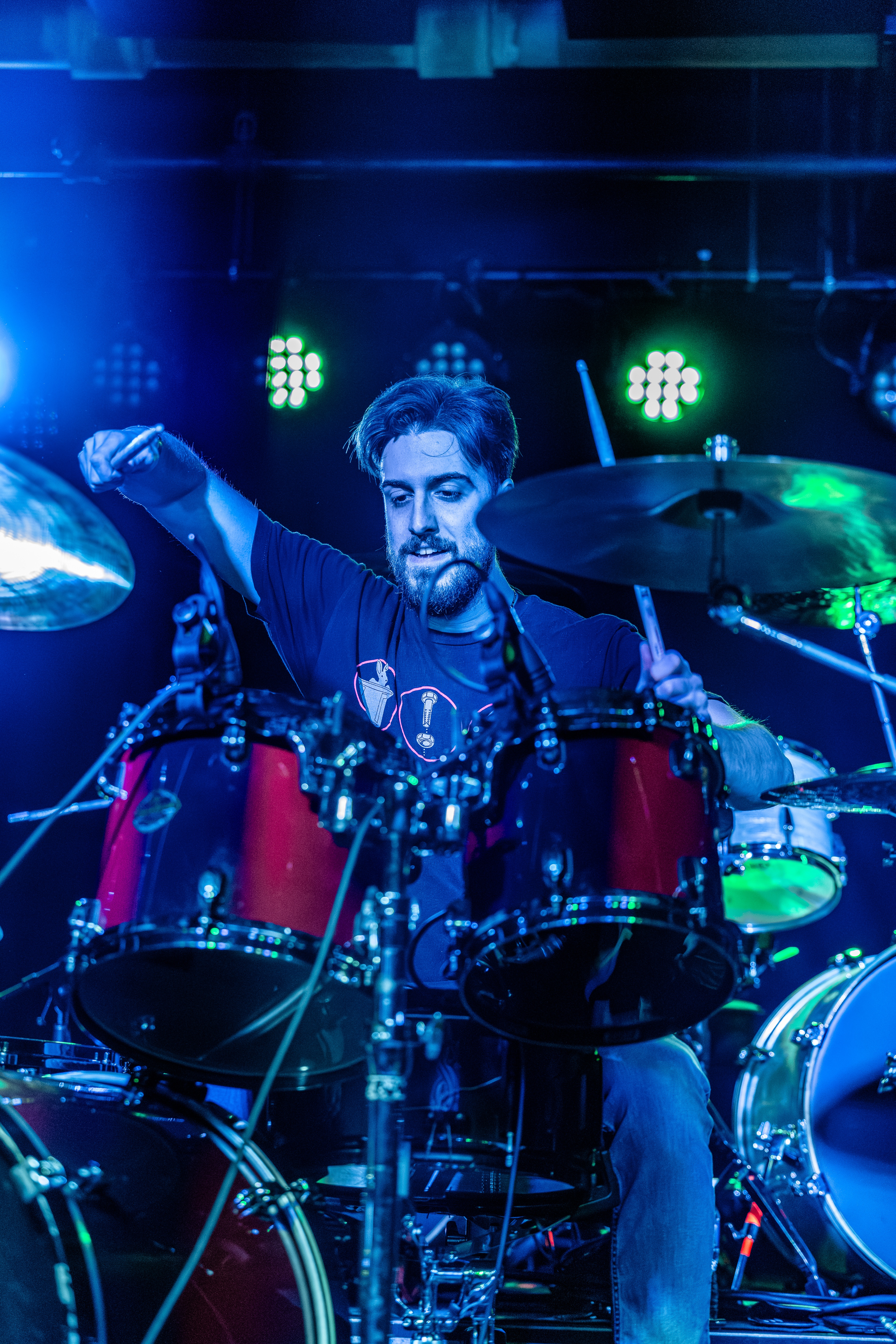 A drummer on stage behind red drums, fist raised, bathed in blue and green stage lighting with drum kit visible.