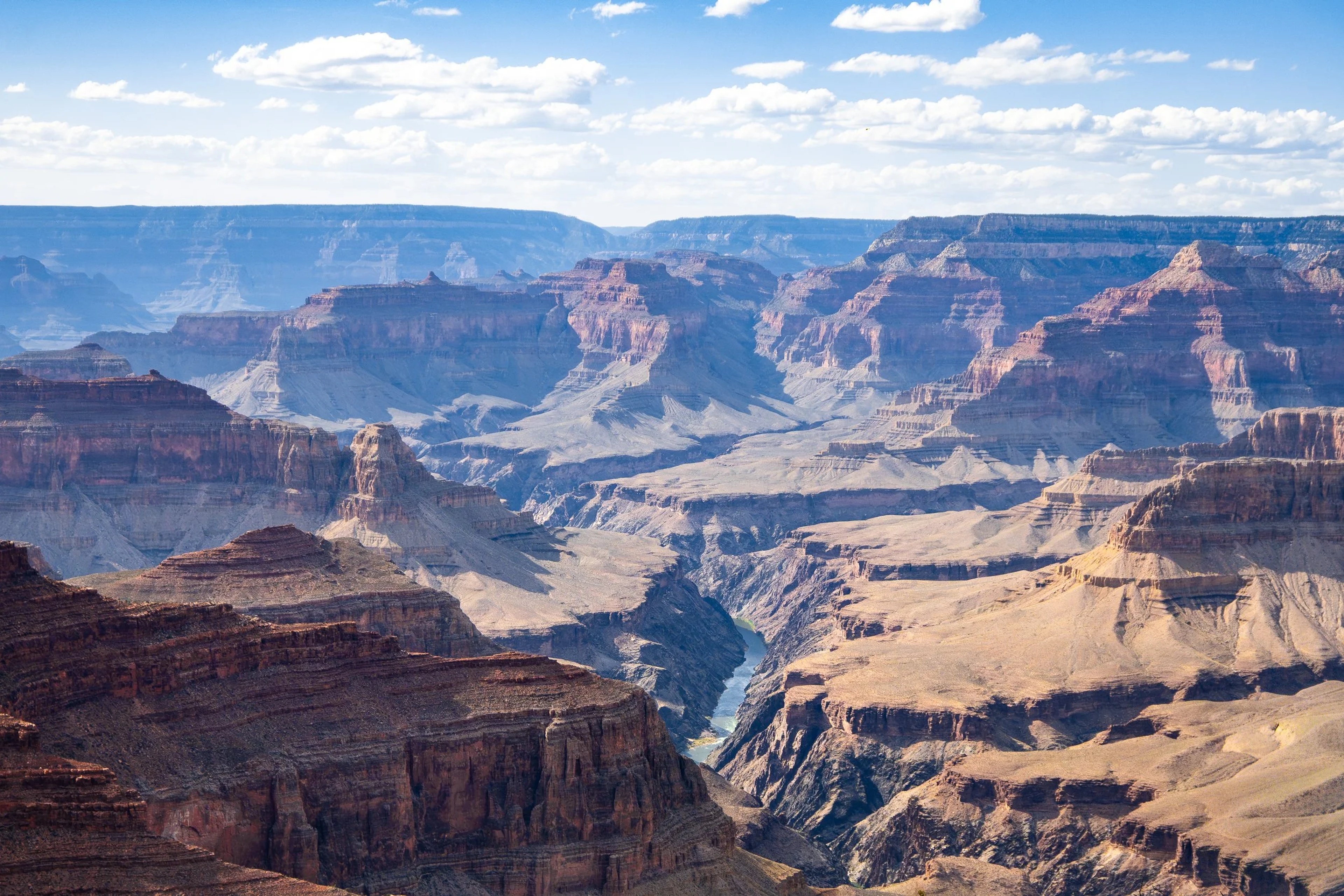 Expansive aerial view of the Grand Canyon showing layered rock formations, the Colorado River winding through the canyon floor, and blue sky with white clouds overhead.