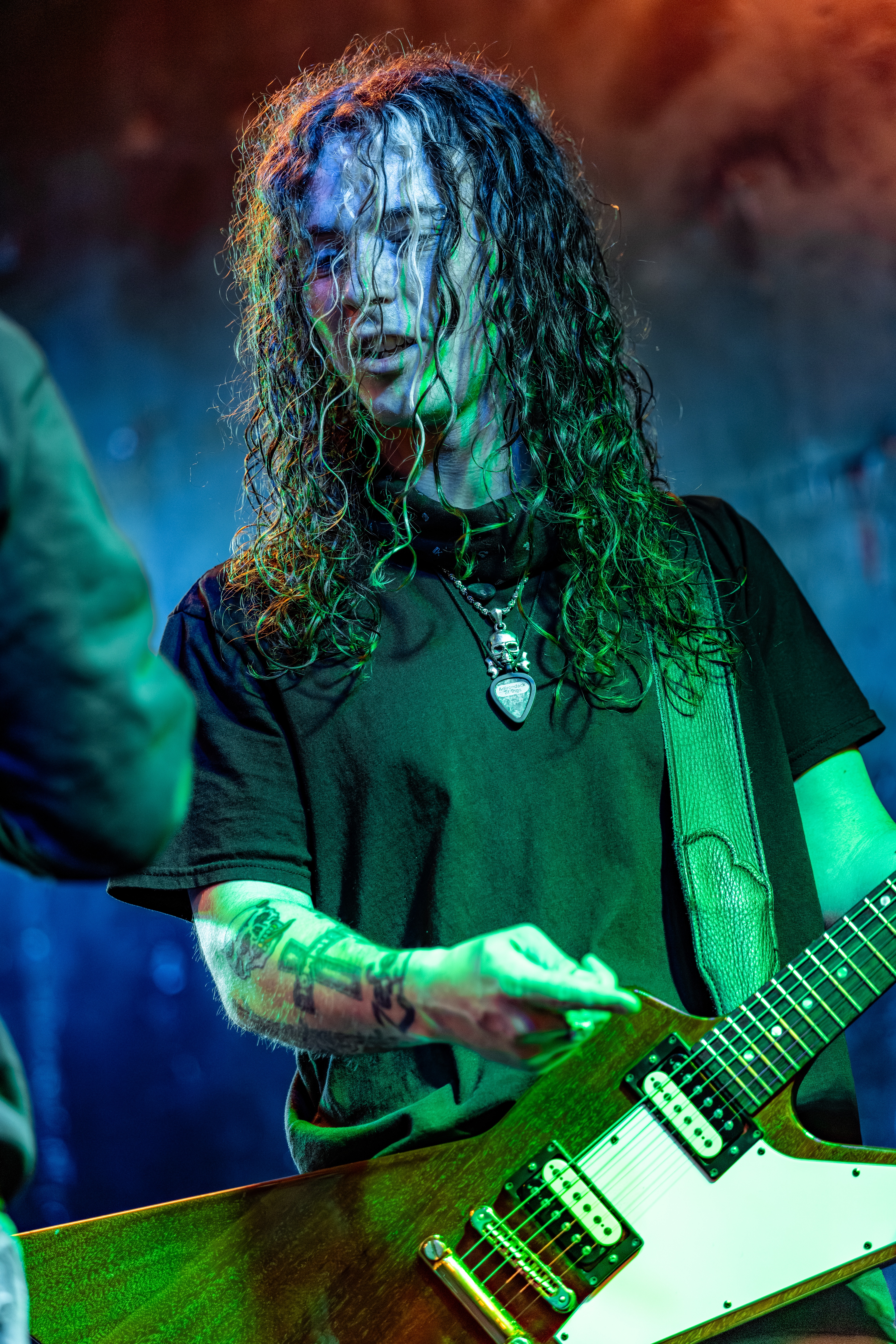 A long-haired musician on stage holding an electric guitar, lit in bright green and blue stage lights with sweat visible on his skin.