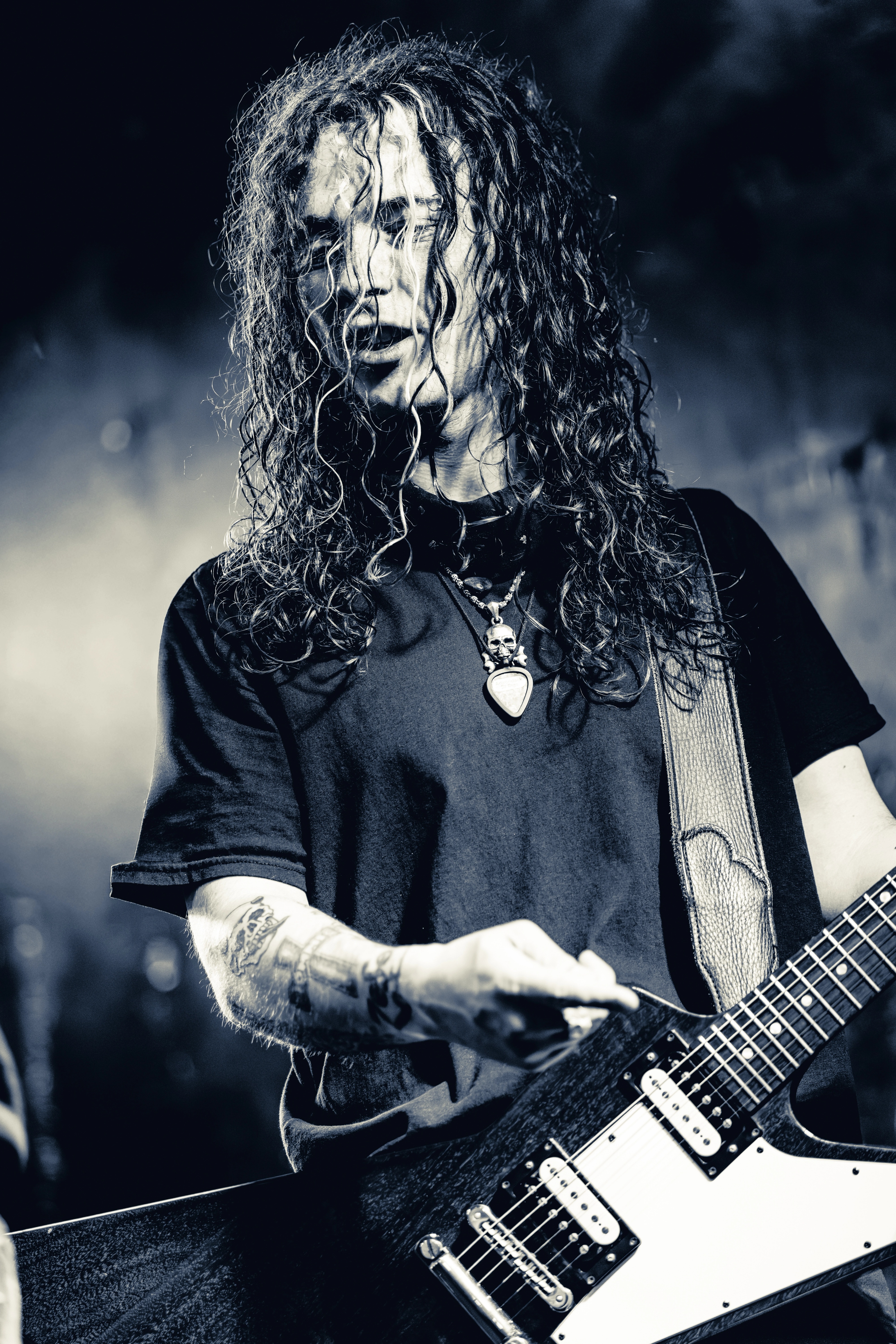 A long-haired musician plays electric guitar on stage, silhouetted against blue lighting and stage smoke, wearing a dark t-shirt and pendant necklace.