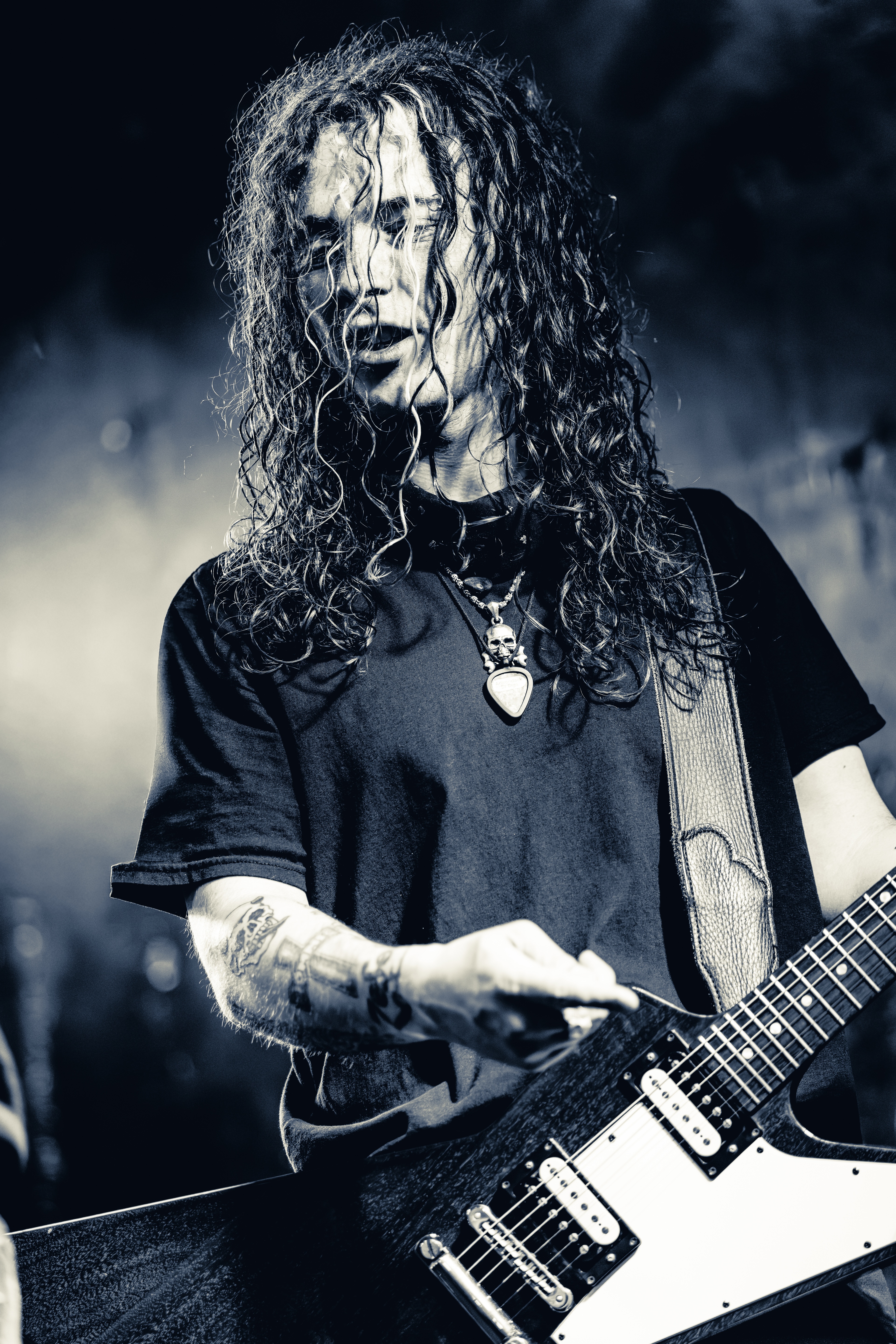 A long-haired musician plays electric guitar on stage, silhouetted against blue lighting and stage smoke, wearing a dark t-shirt and pendant necklace.