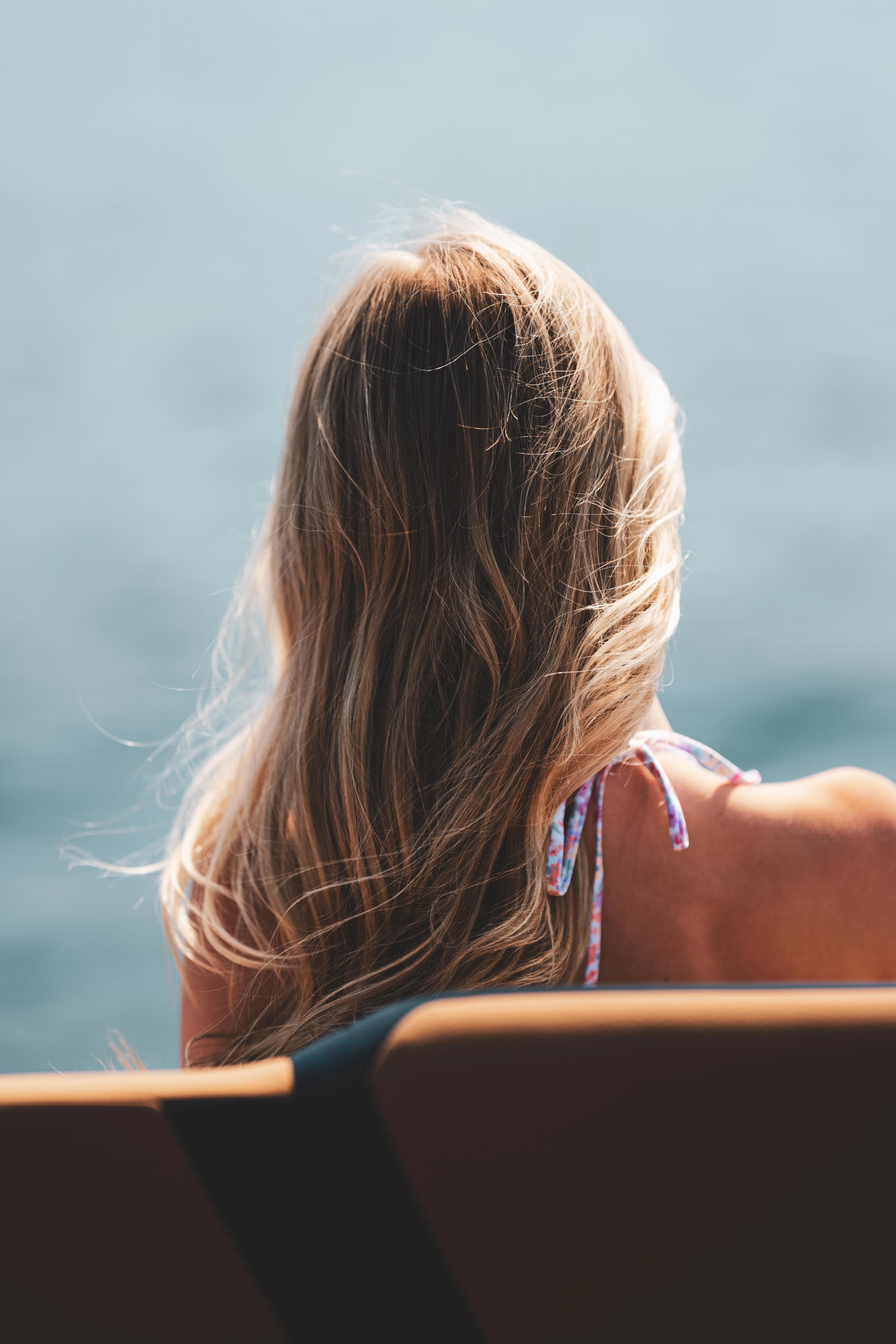 A woman with long brown hair seen from behind, gazing toward a blue lake under clear sky at an overlook.