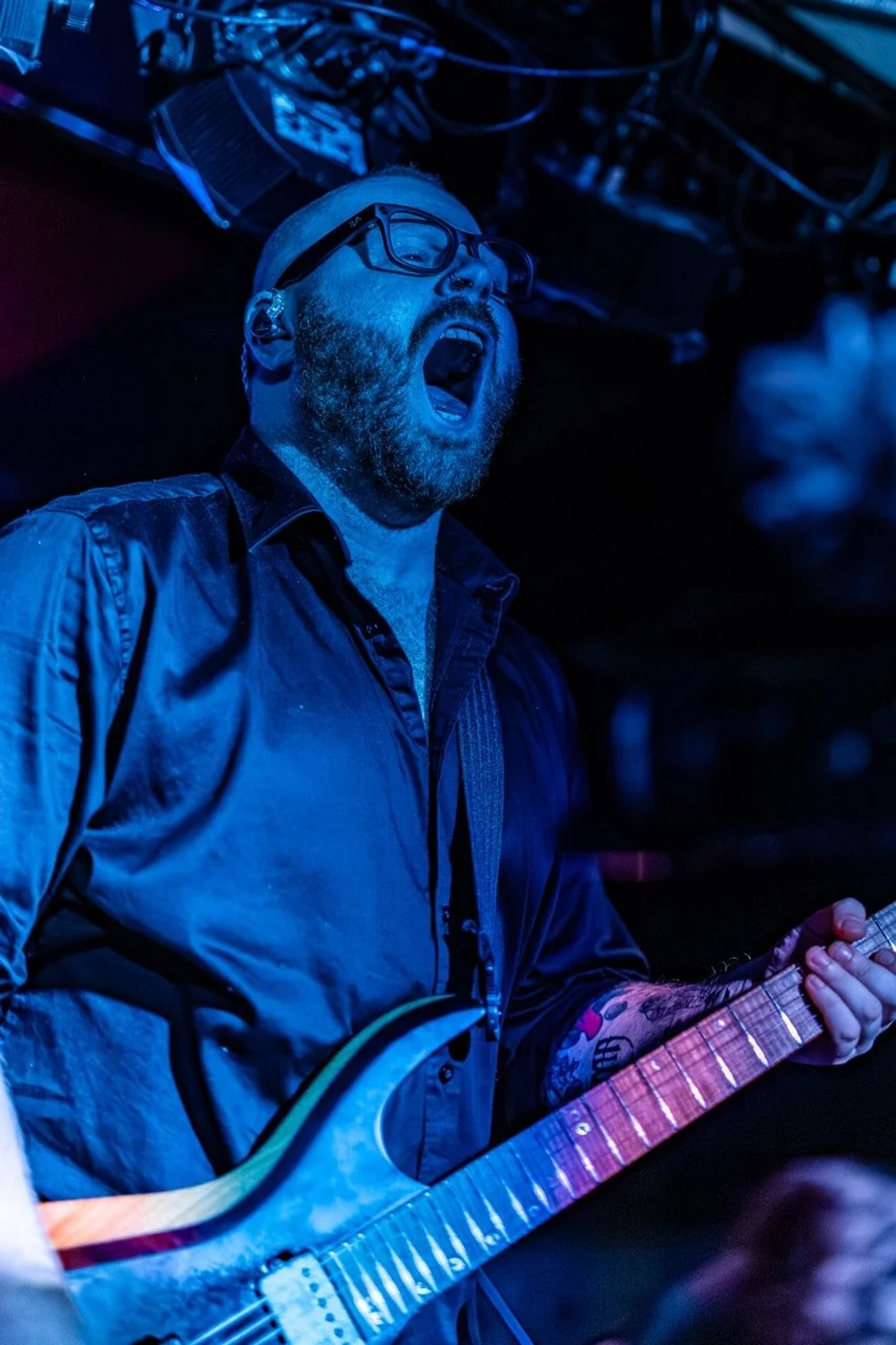 A bearded guitarist in glasses performs on stage under blue stage lighting, holding an electric guitar while singing intensely.
