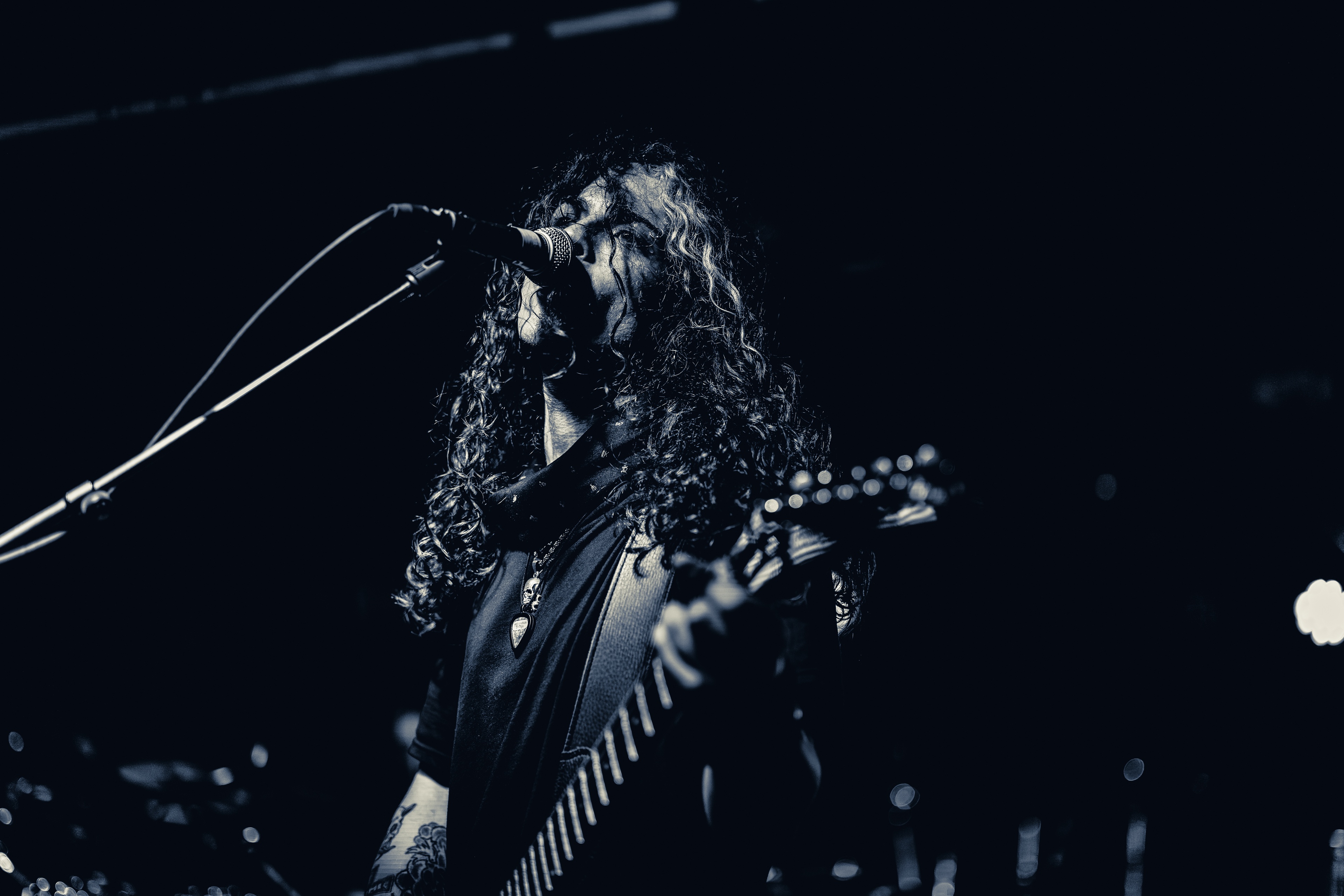 A long-haired musician performs at a microphone while playing an electric guitar, lit dramatically against a dark stage with ambient bokeh lights.