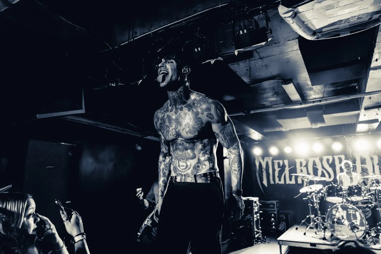 A shirtless, heavily tattooed male vocalist performs on stage at Empire Live, mouth open mid-scream, with drum kit and band backdrop visible behind him.
