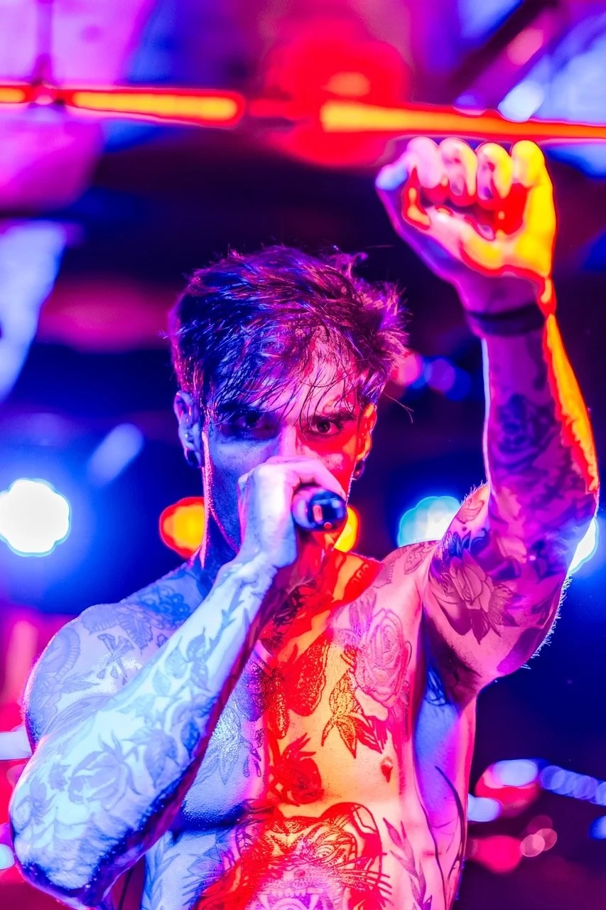 A shirtless tattooed vocalist performs into a microphone, fist raised, bathed in vibrant neon pink, blue, and yellow stage lighting.