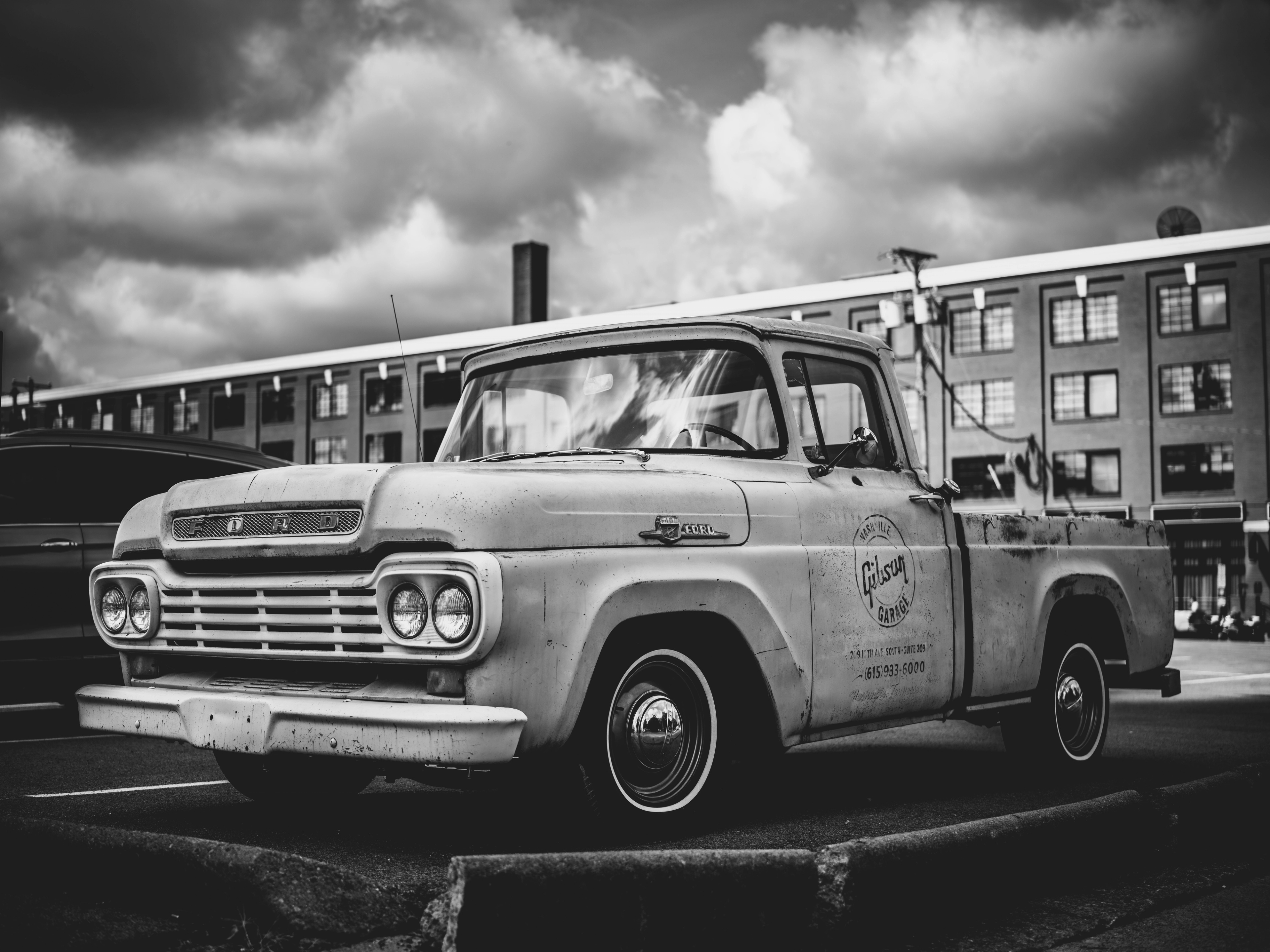 A weathered vintage Ford pickup truck with patina paint and chrome details parked in front of an industrial brick building under dramatic cloudy sky.