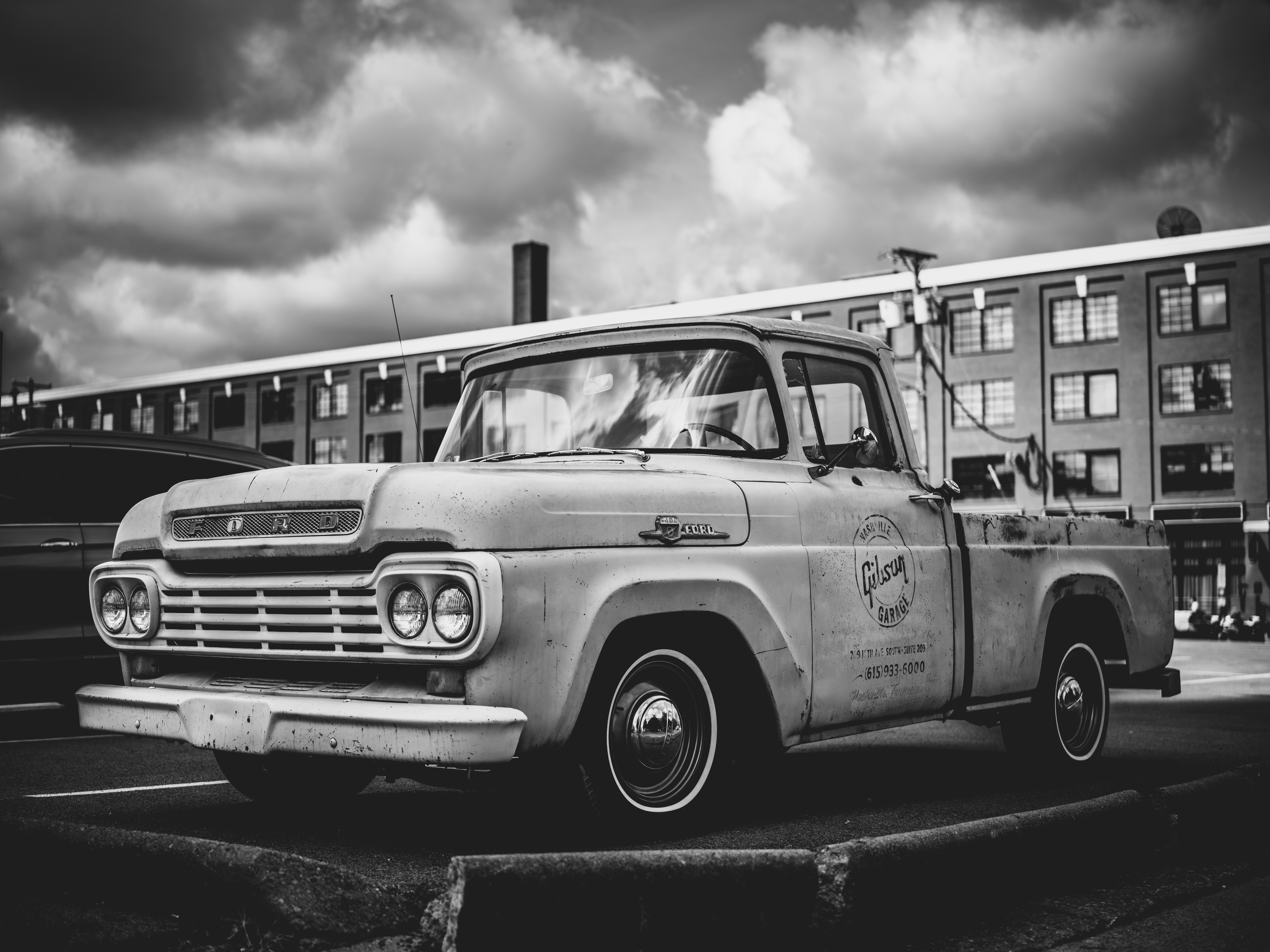 A weathered vintage Ford pickup truck with patina paint and chrome details parked in front of an industrial brick building under dramatic cloudy sky.