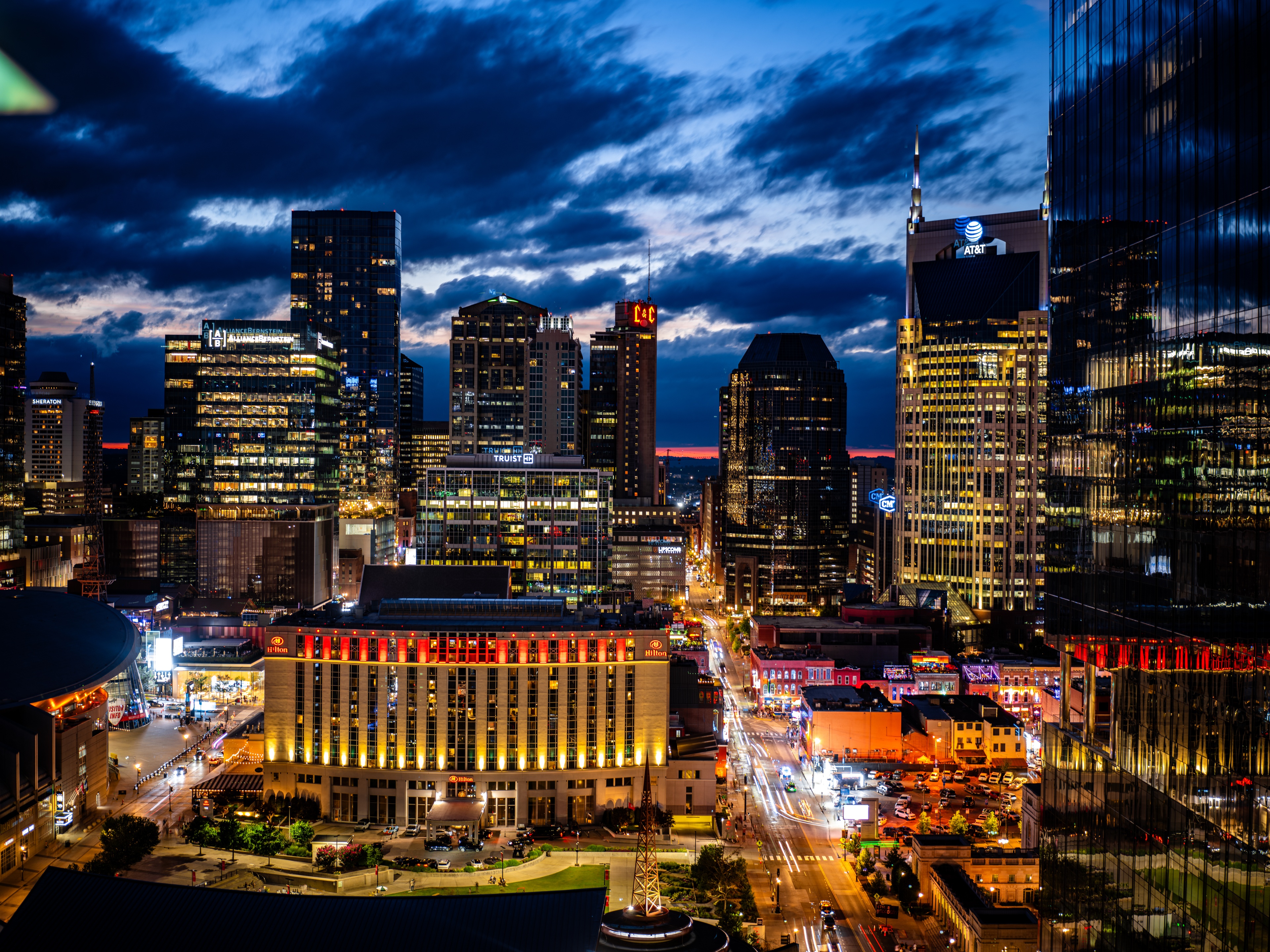 A city skyline at dusk illuminated by golden building lights against a dramatic blue and purple stormy sky with clouds.