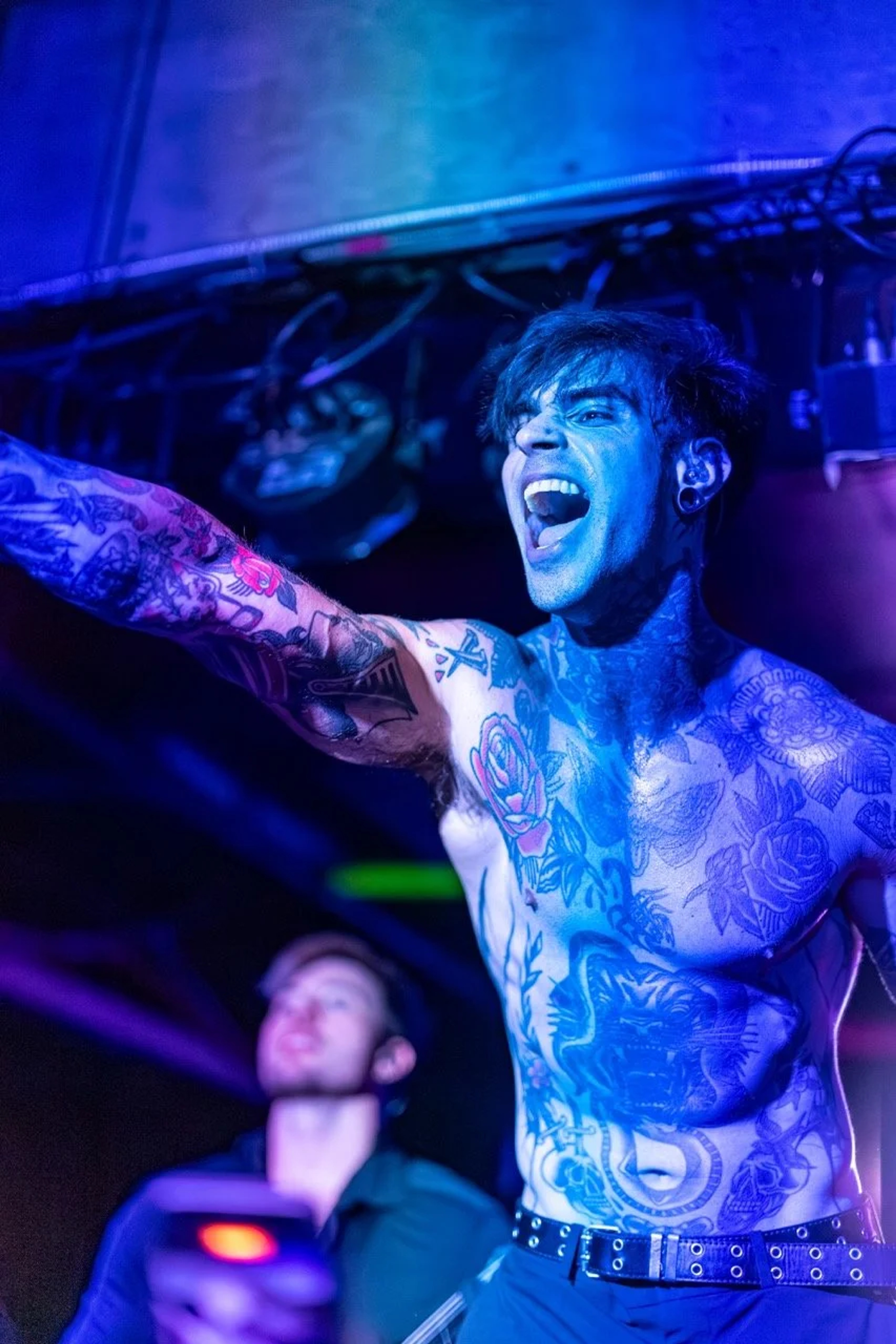 A shirtless tattooed musician on stage at Empire Live Underground, mouth open mid-performance, arm raised, bathed in blue and purple stage lighting.