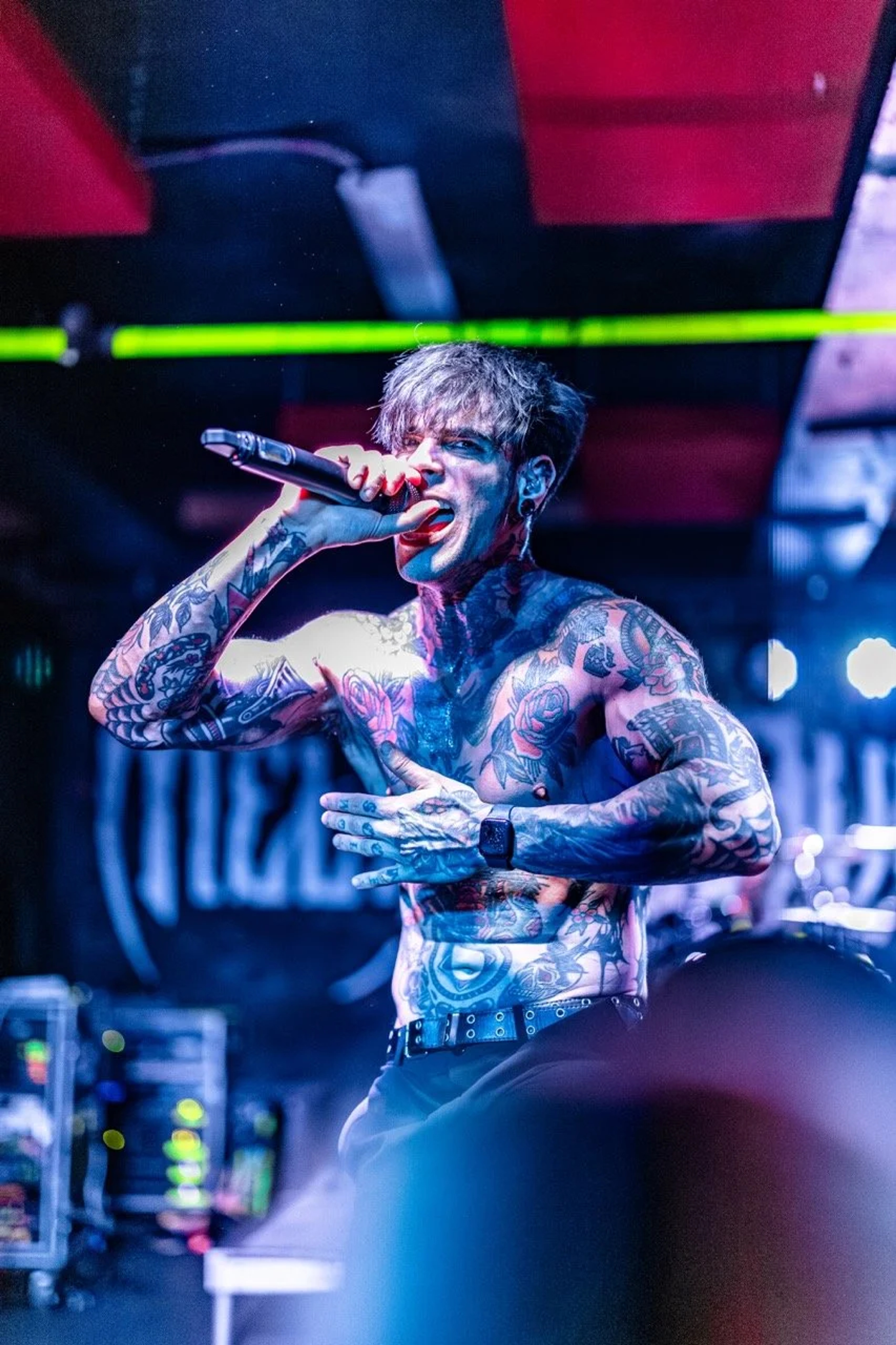 A shirtless tattooed singer grips a microphone on stage, backlit by neon red and blue lighting at an underground venue.