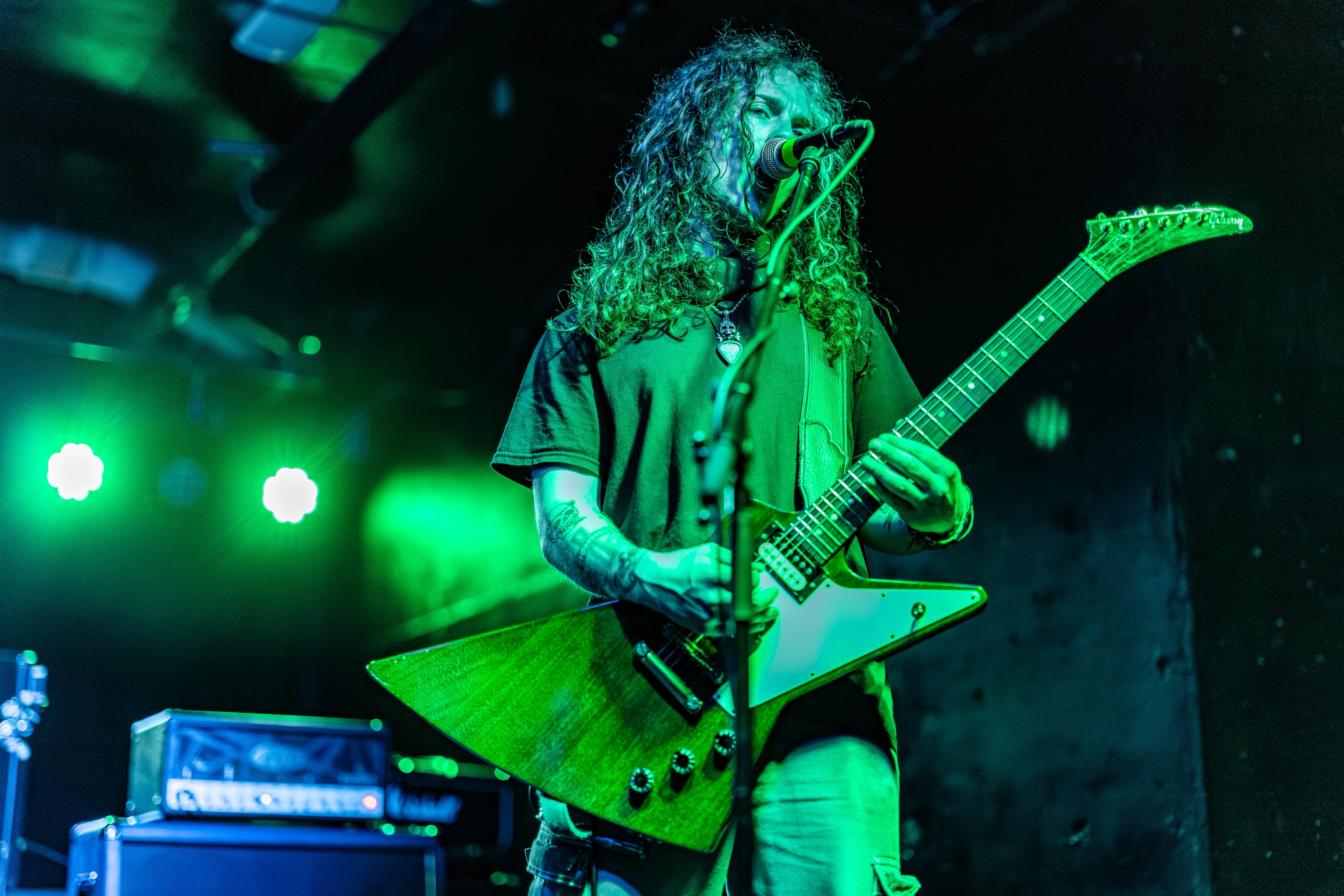 A long-haired guitarist performs on stage under green stage lighting, playing an angular electric guitar while singing into a microphone.