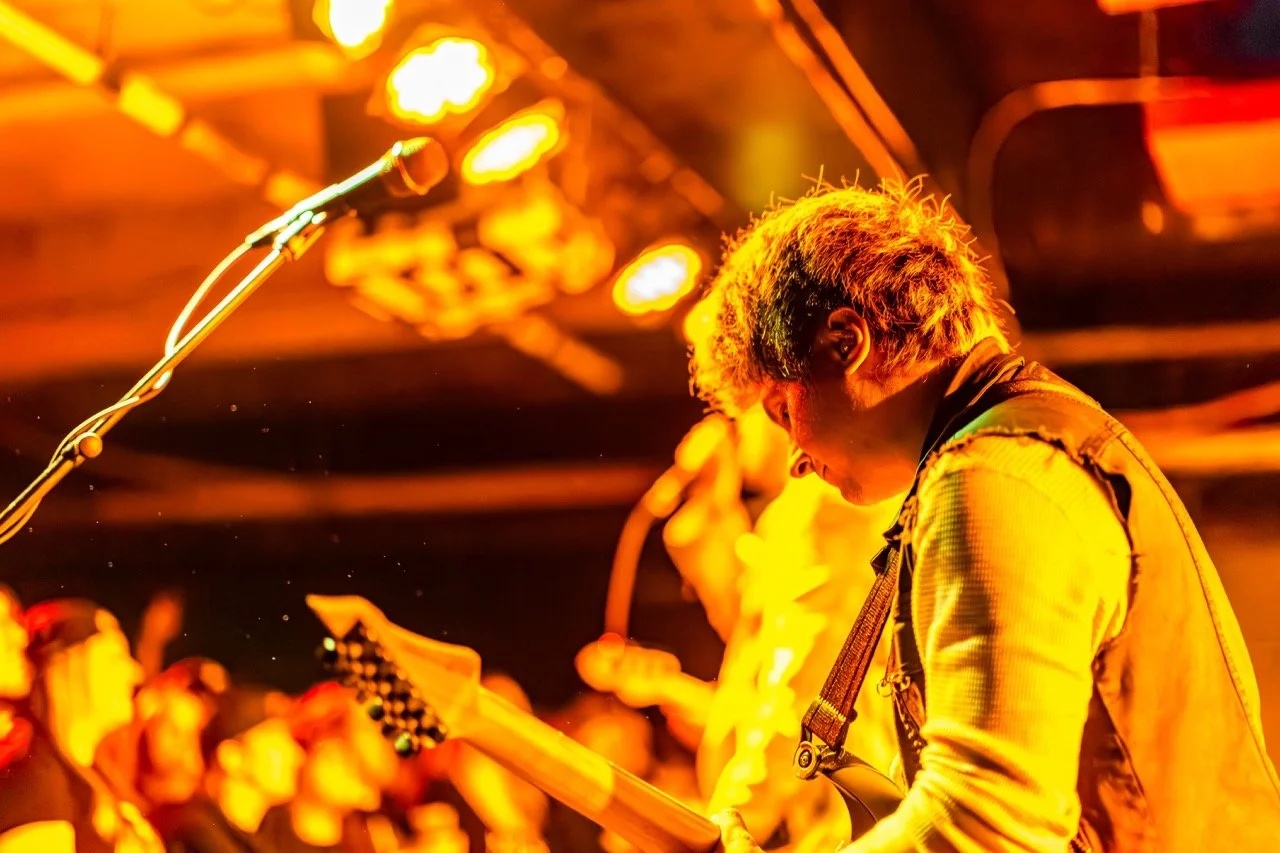 A guitarist performs on stage under warm orange stage lighting, singing into a microphone with intensity at Empire Live Underground.