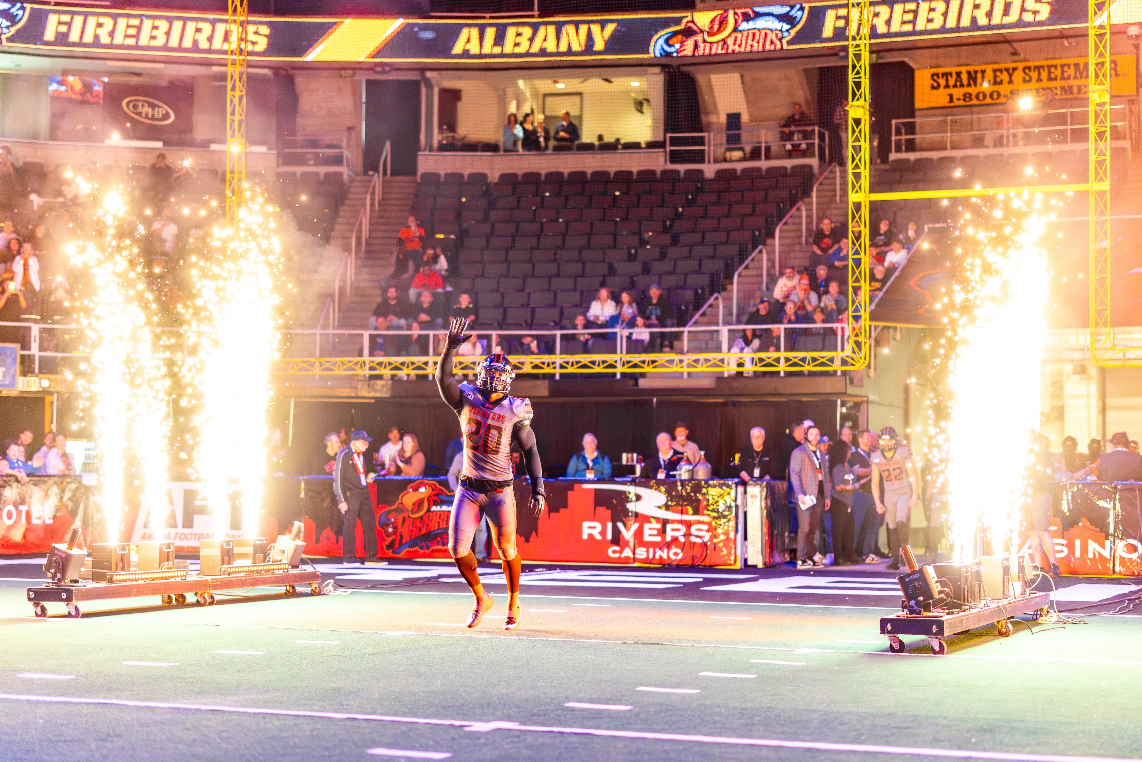 A performer in team colors enters an indoor arena through pyrotechnic flames, with crowd seating and sponsor signage visible in the background.