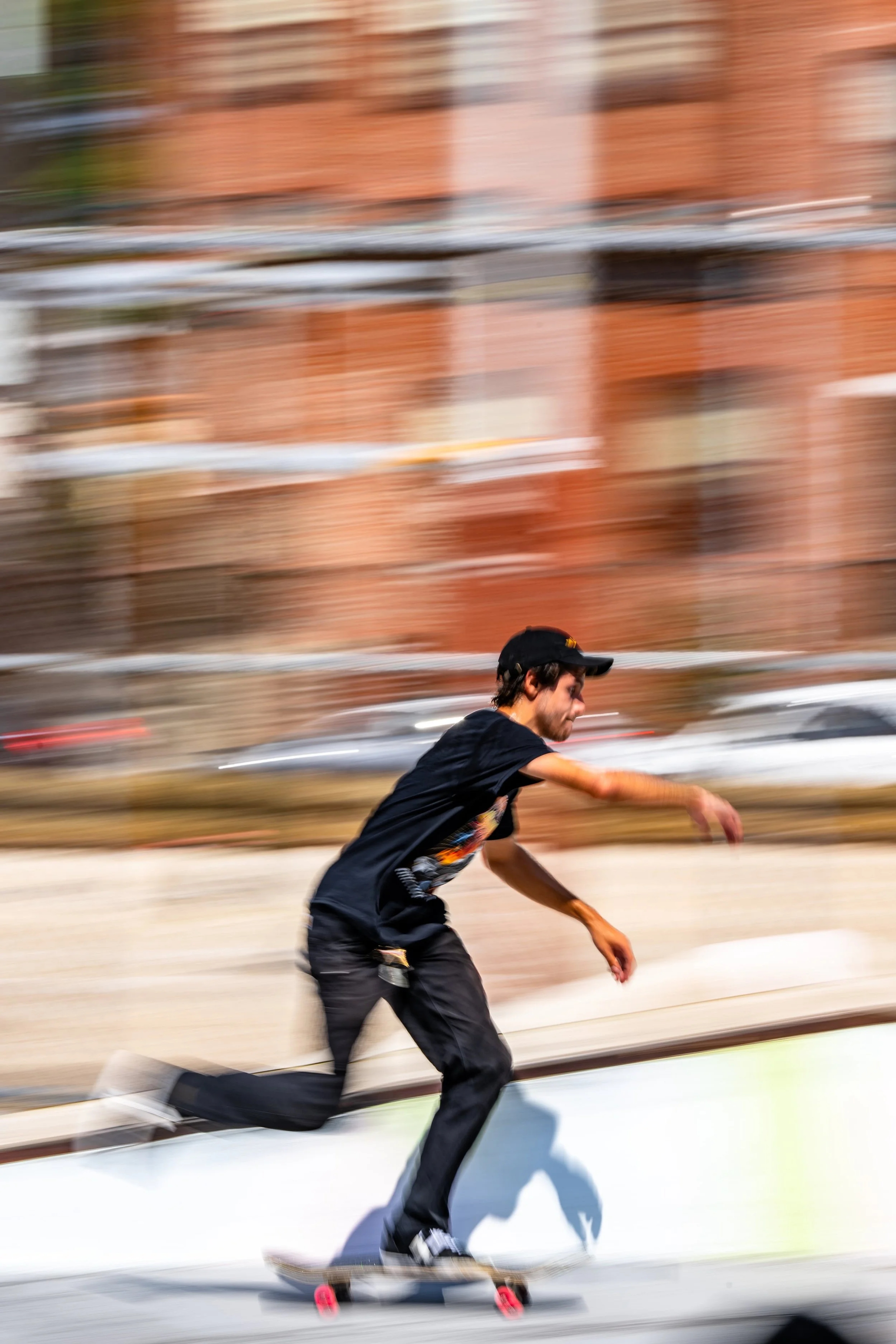 A skateboarder in black shirt and cap rides a skateboard down a city street, with a brick building and blurred motion in the background.