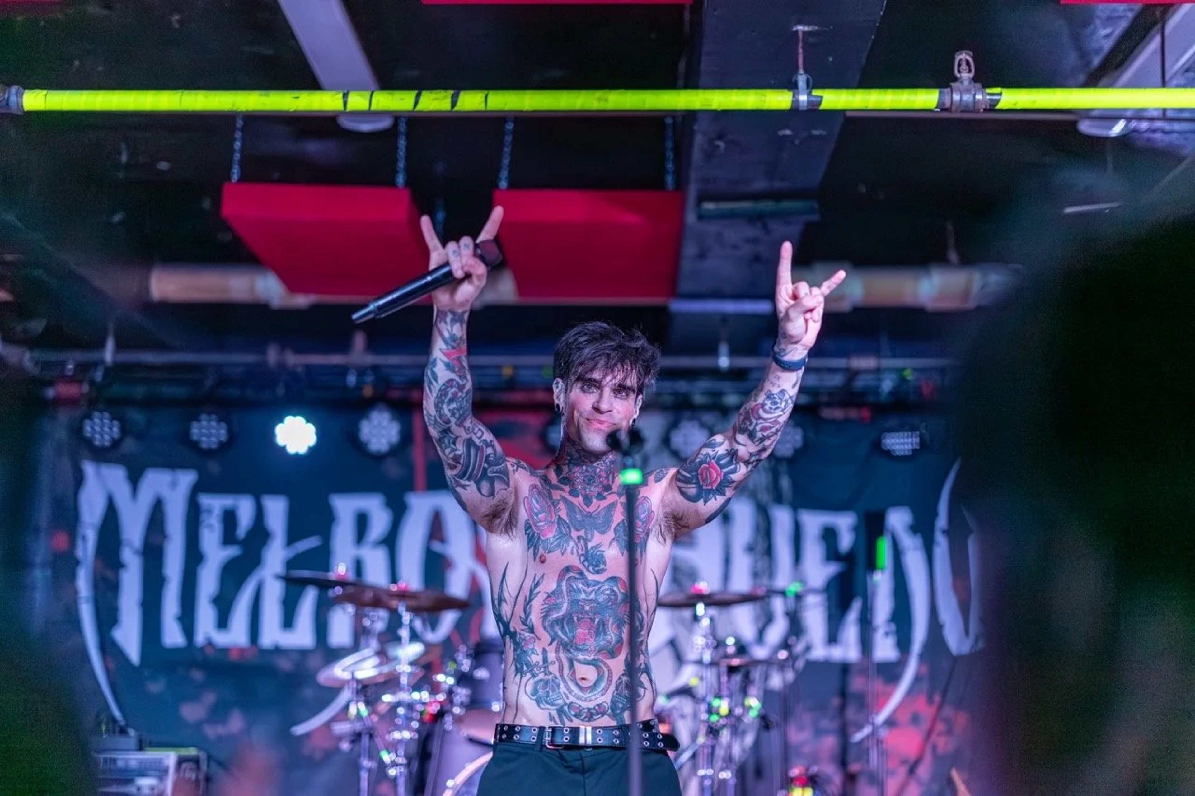 A shirtless, tattooed male vocalist on stage holding a microphone with both arms raised in a rock salute, illuminated by vibrant stage lighting and geometric green rigging overhead.