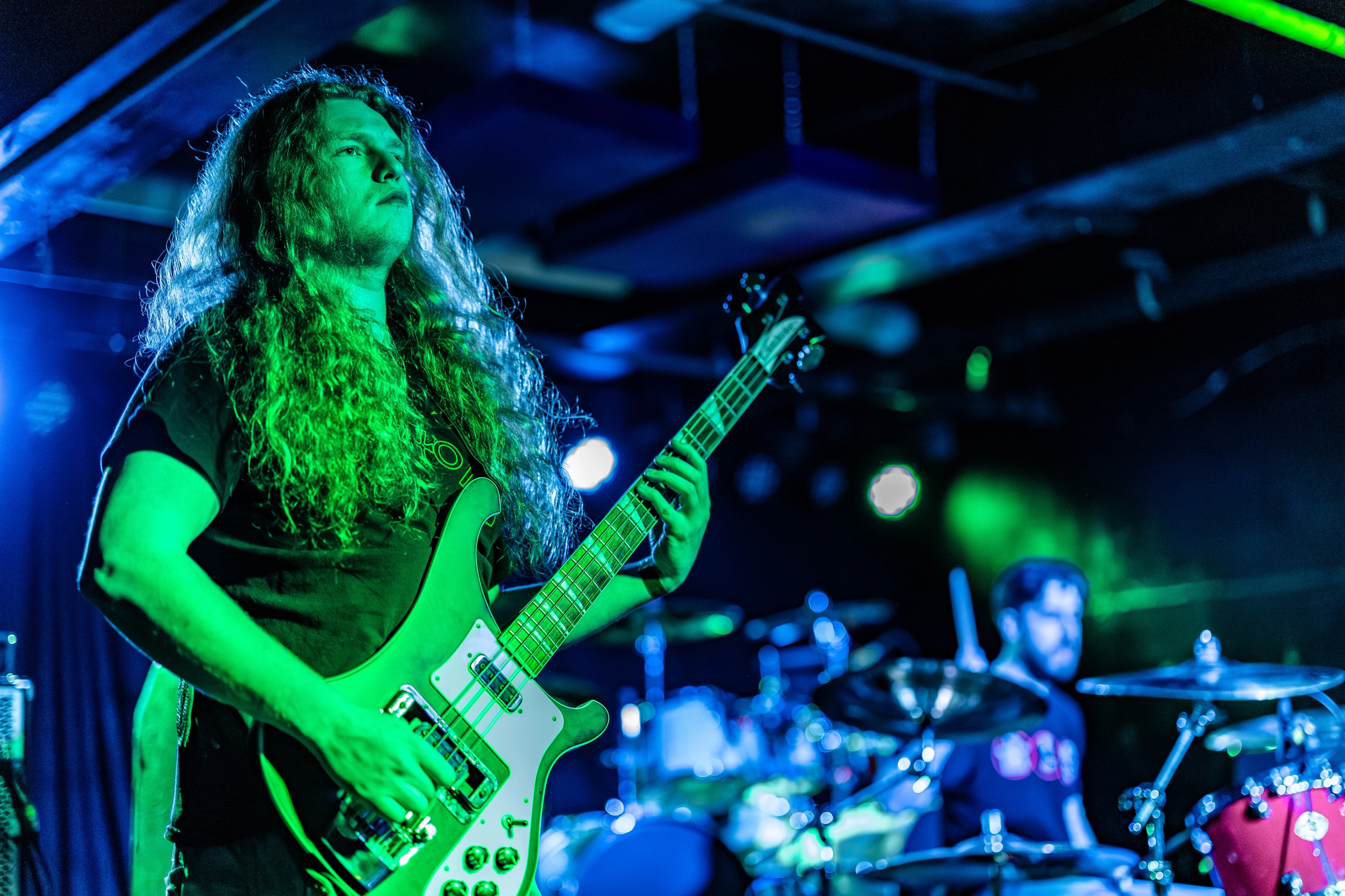 A long-haired guitarist on stage holding an electric guitar, bathed in green stage lighting with blue lights and drums visible in the background.
