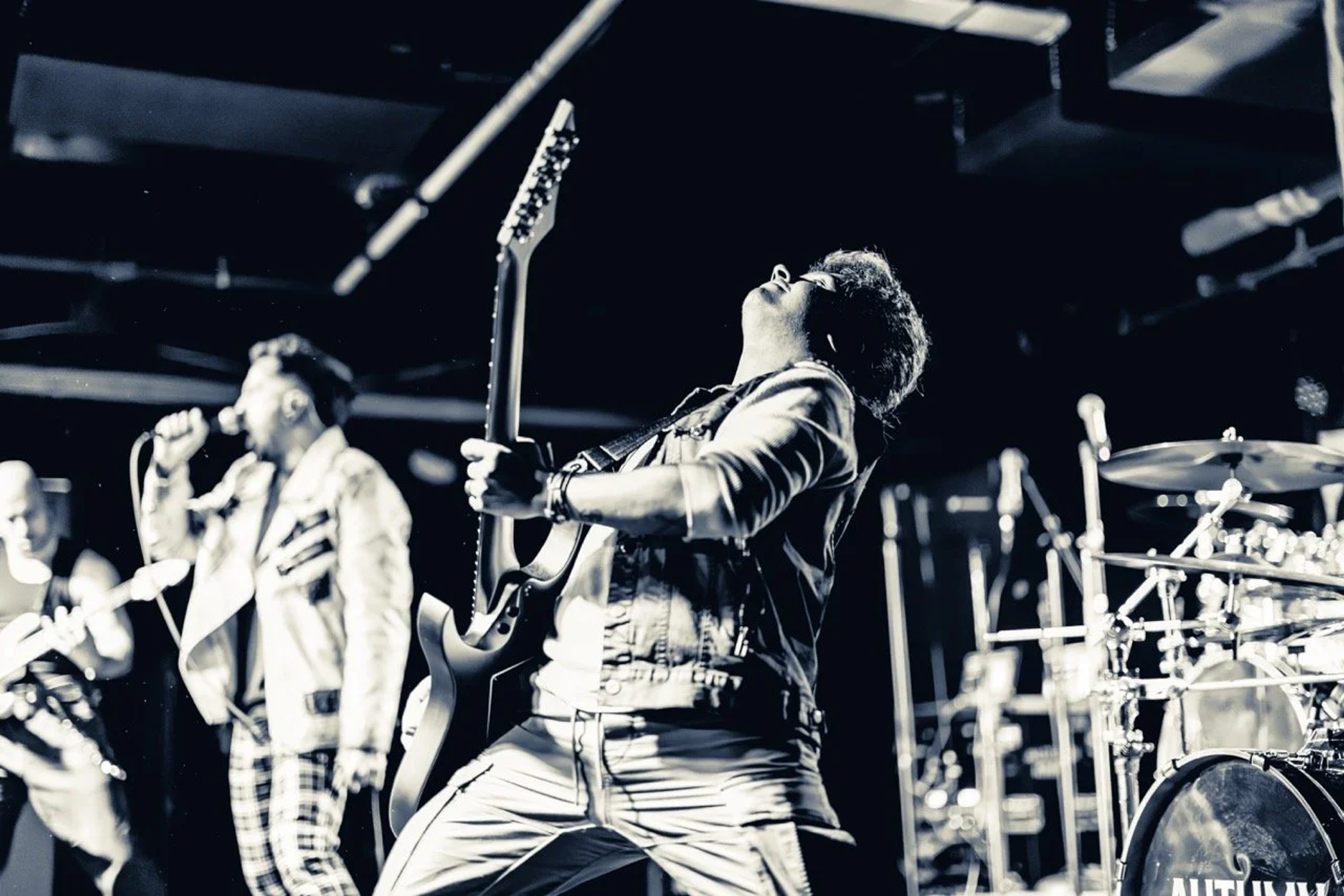 A guitarist throws his head back mid-solo on stage at Empire Live Underground, guitar raised, with a vocalist and drummer visible in the background under harsh stage lighting.