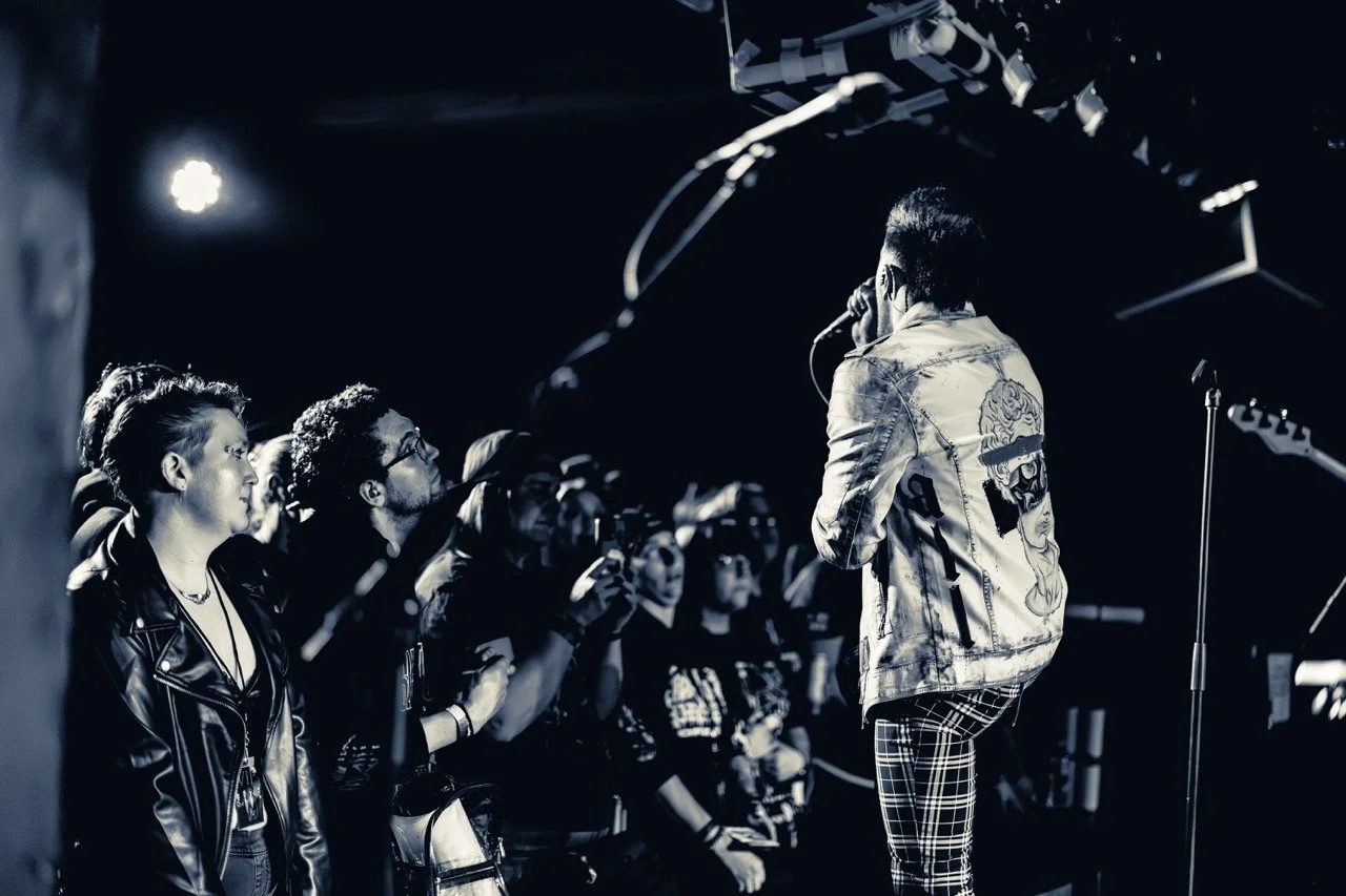 A shirtless vocalist on stage holding a microphone, surrounded by audience members in a crowded pit, lit by overhead stage lights in a dark venue.