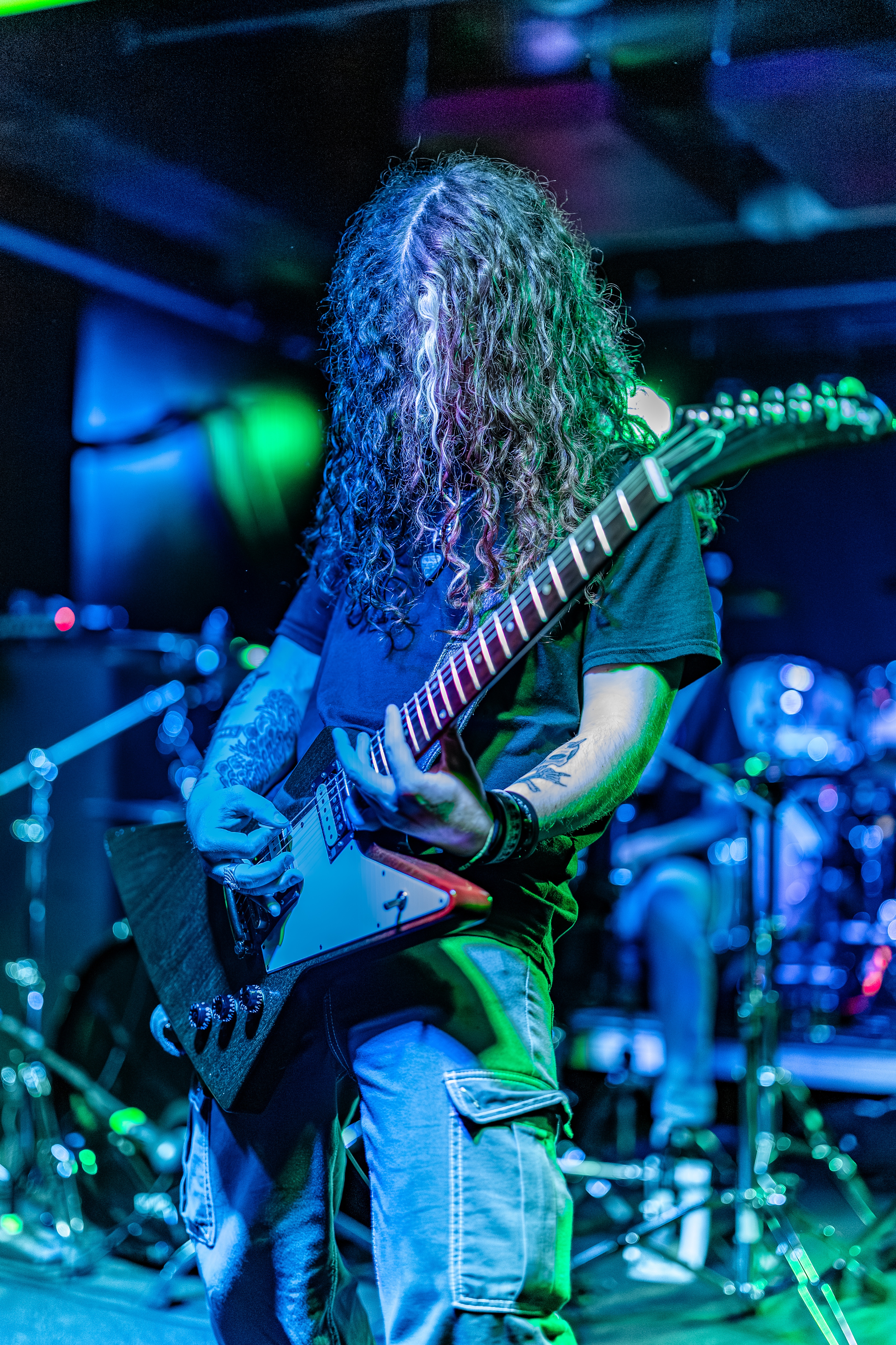 A long-haired guitarist in mid-performance on stage, illuminated by blue and green stage lighting, playing an electric guitar with intensity.