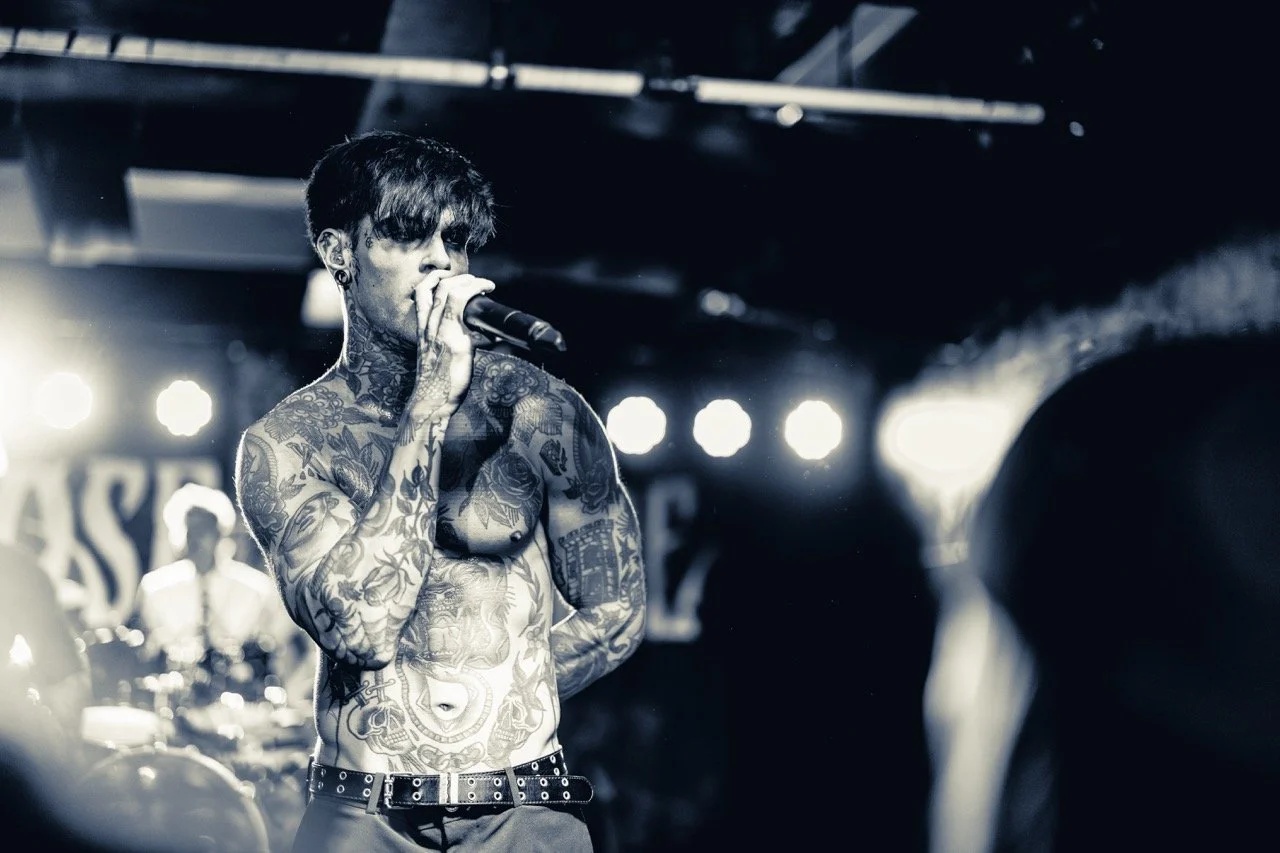 A tattooed singer performs on stage at Empire Live Underground, holding a microphone to his mouth with stage lights and blurred crowd visible behind him.