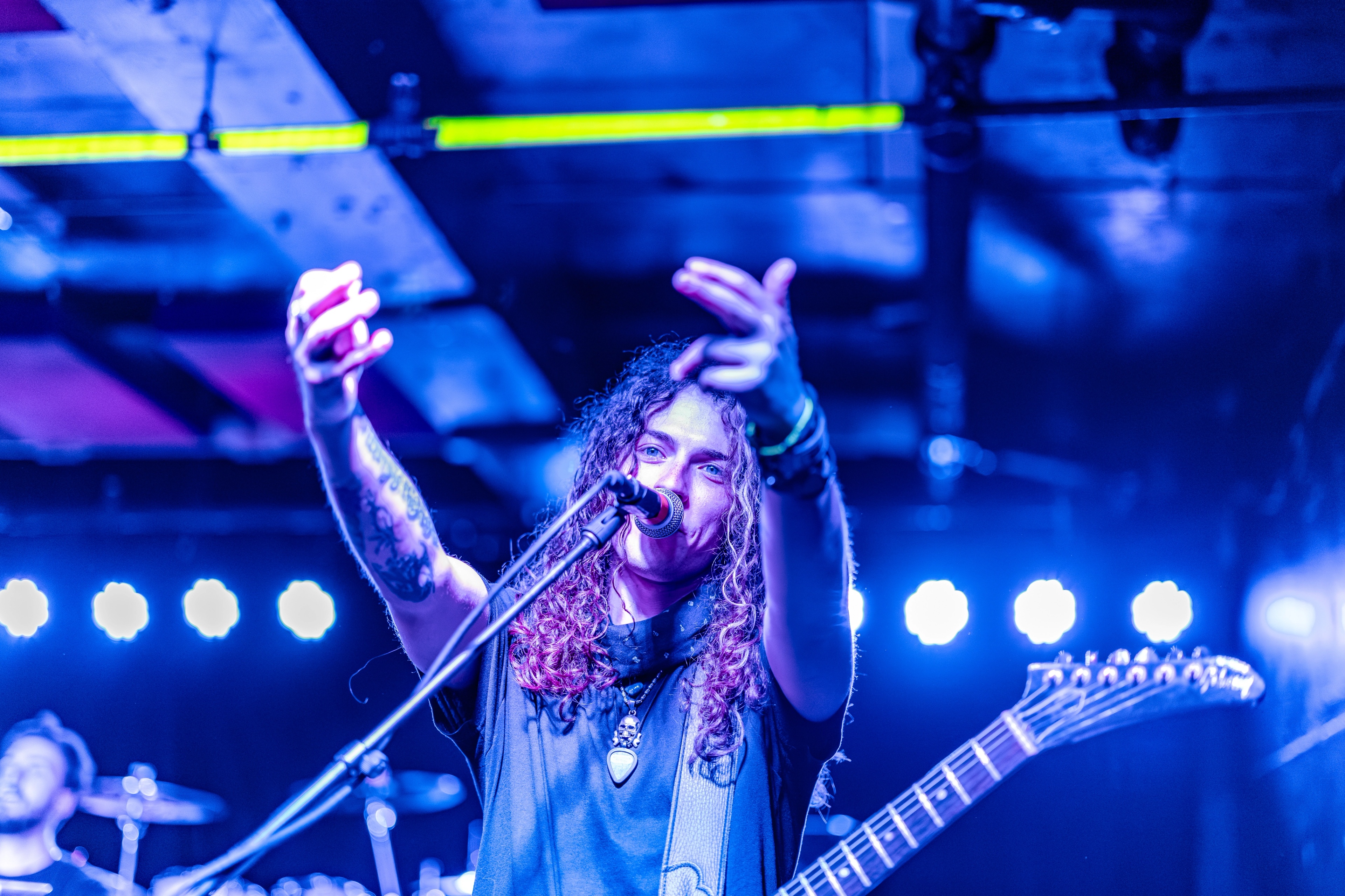 A long-haired vocalist on stage with microphone, arms raised, surrounded by blue and neon stage lighting and band equipment.