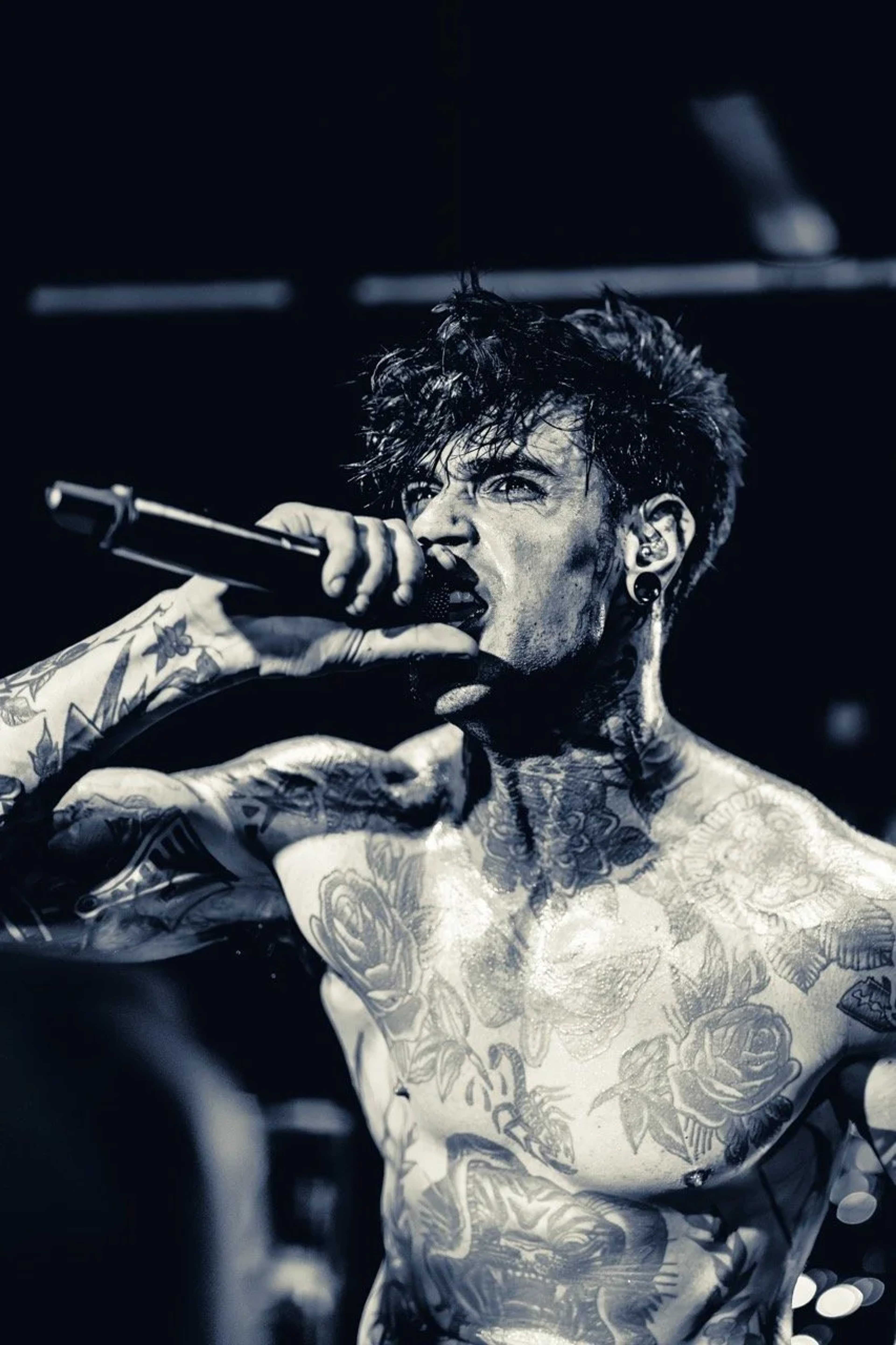 A shirtless tattooed male vocalist performs on stage holding a microphone, captured in dramatic black and white lighting against a dark background.