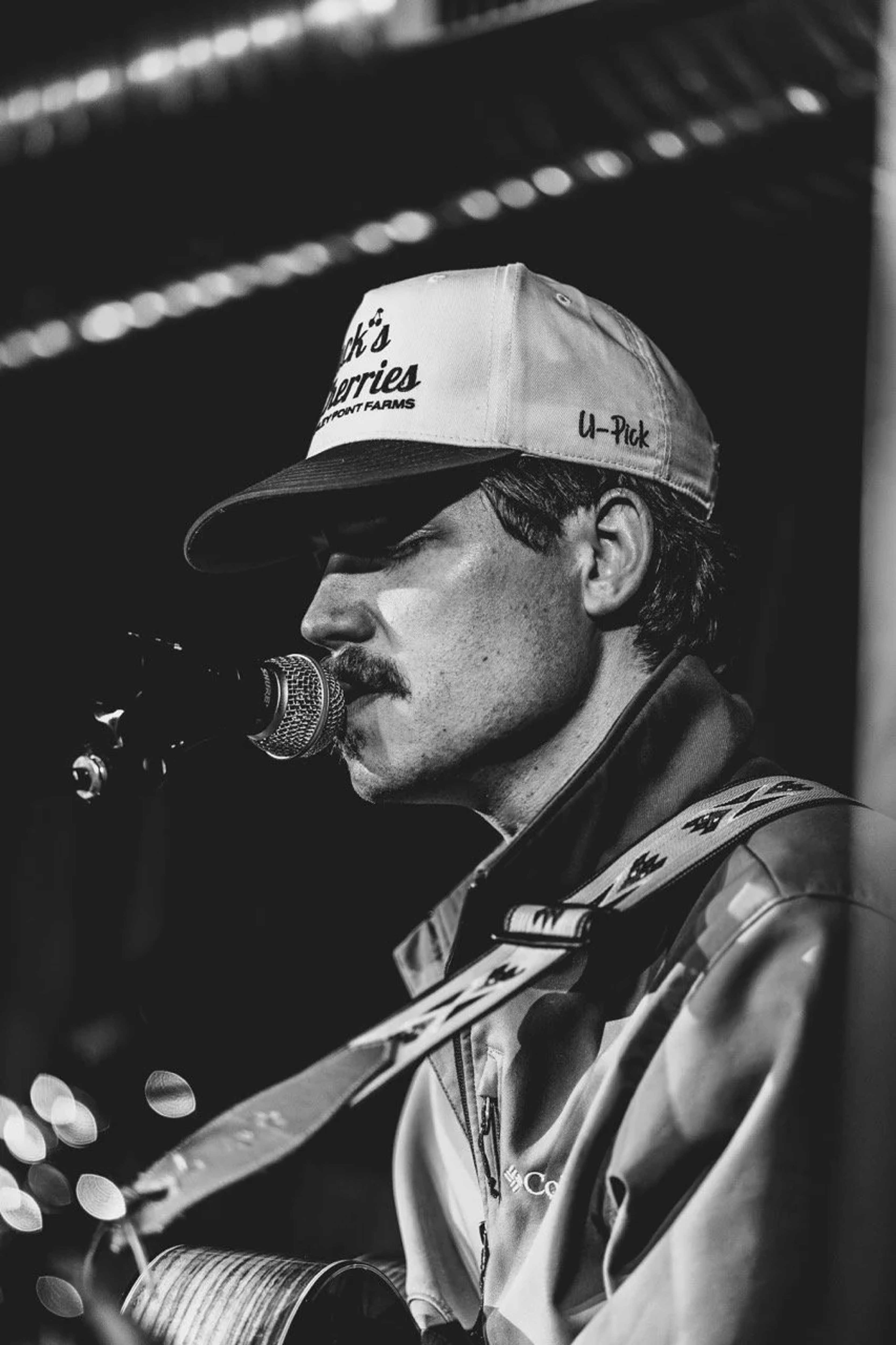 A bearded musician singing into a microphone, wearing a U-Pick farm cap and patterned jacket under stage lights.