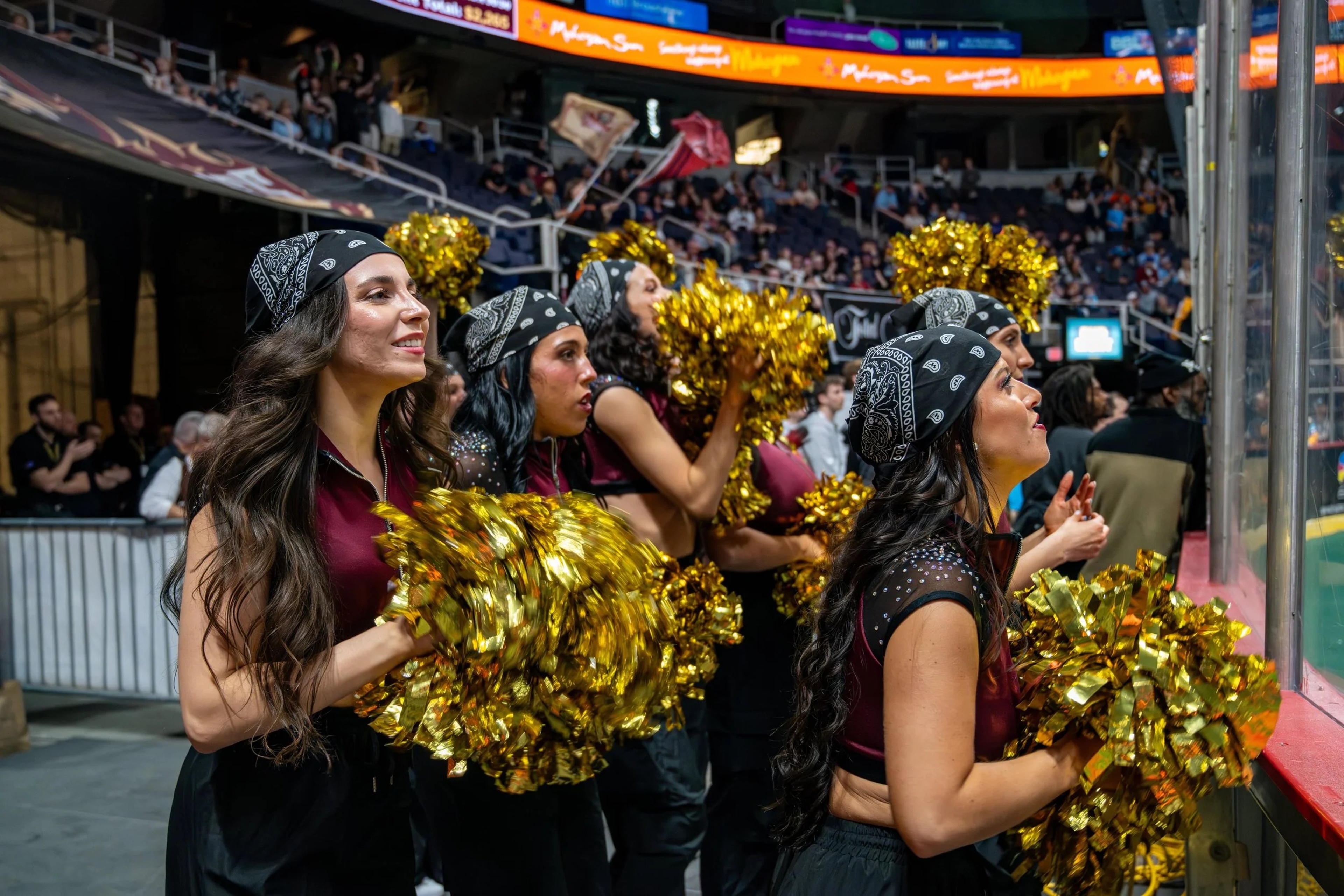 Cheerleaders in black and maroon uniforms holding gold pom-poms perform on the arena floor during a game at MVP Arena, with crowds visible in the stands.