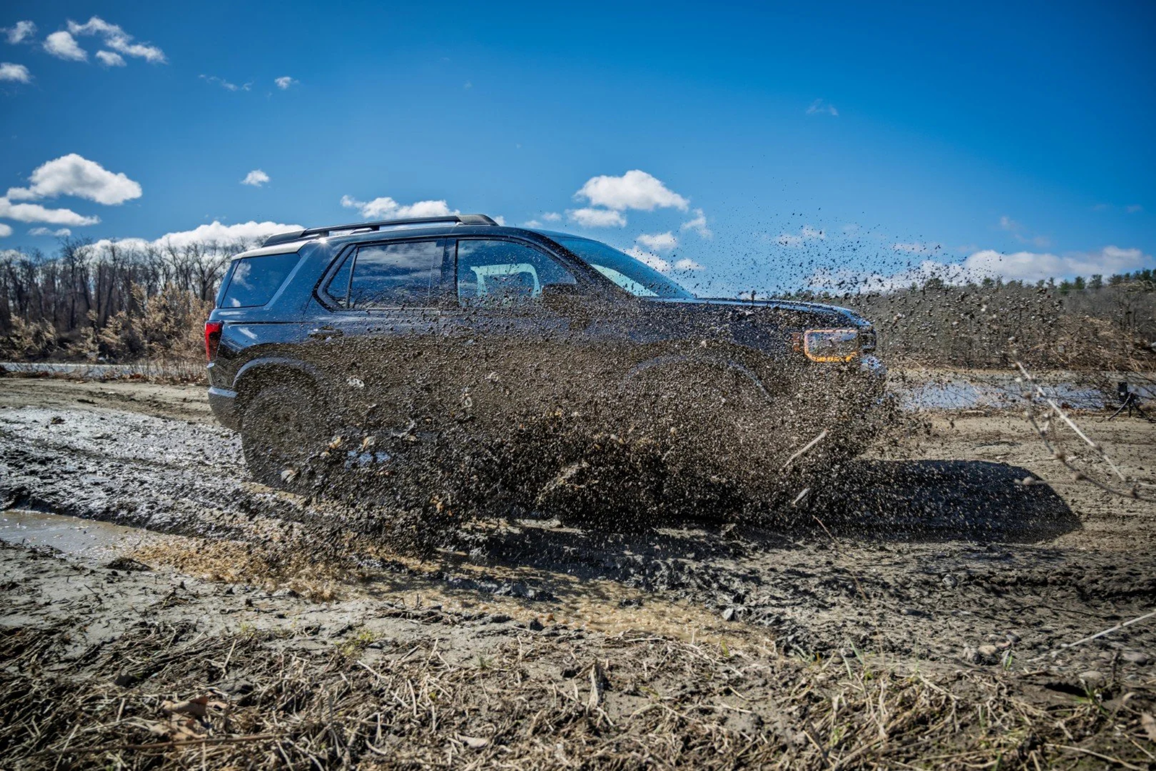 A blue SUV launches through a mud pit, spraying earth and water across the frame against a clear blue sky and forested background.