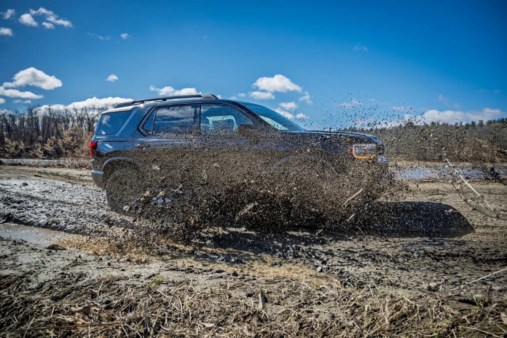 A blue SUV launches through a mud pit, spraying earth and water across the frame against a clear blue sky and forested background.