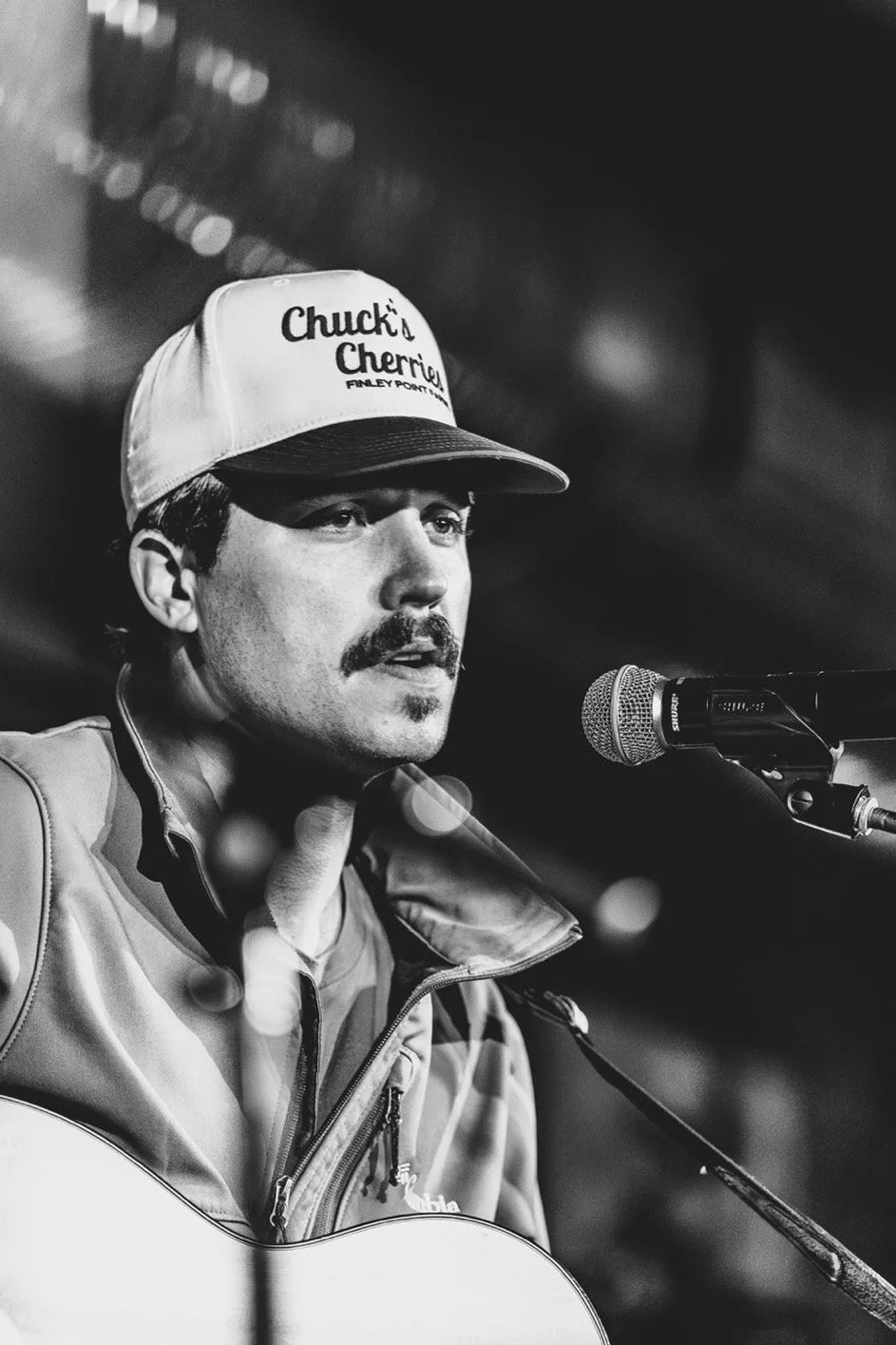 A musician wearing a Chuck's Cherries cap and jacket speaks into a microphone on stage, captured in black and white profile lighting.