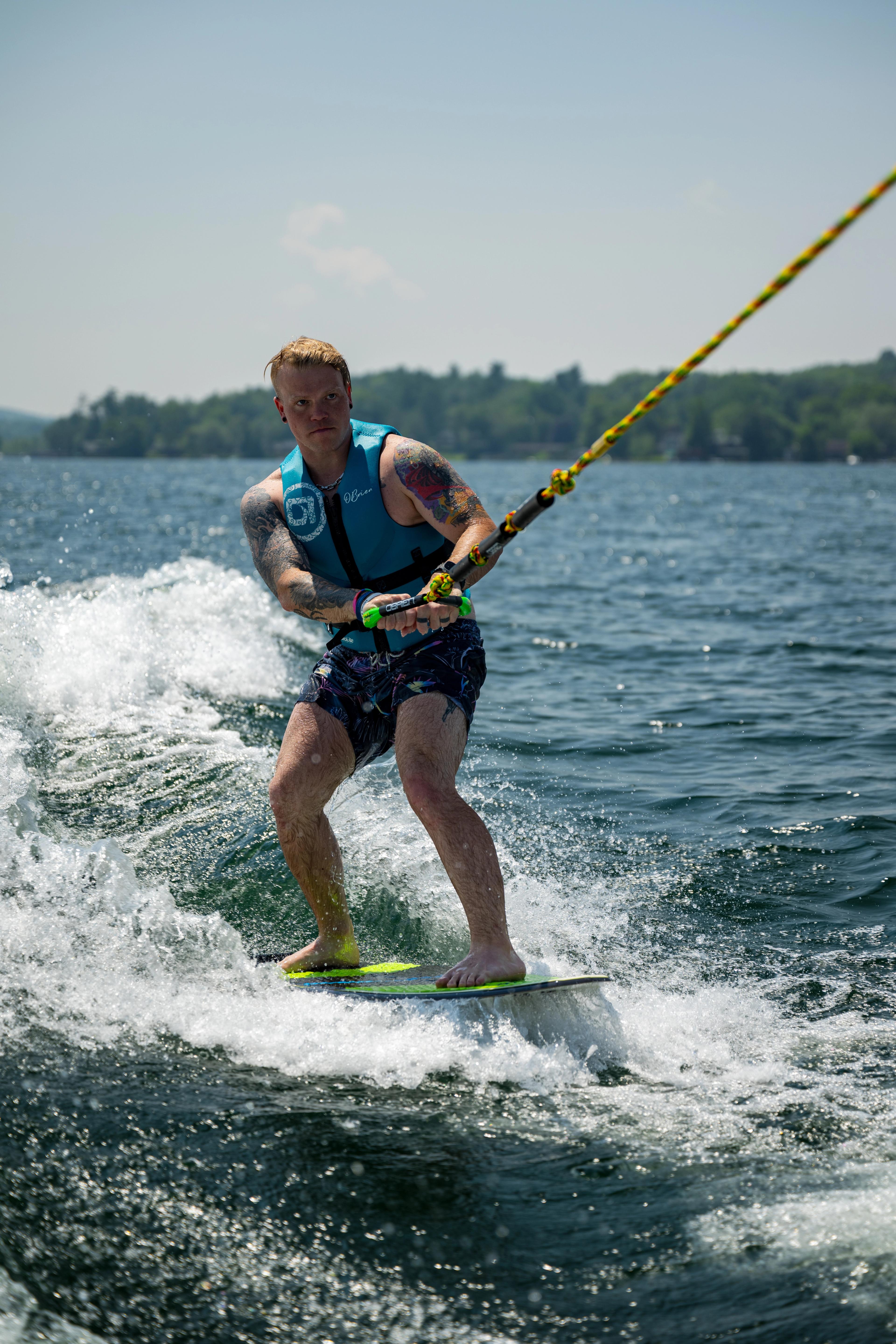 A male wakeboarder on a yellow board being towed by rope across choppy lake water, performing on a sunny day with forested shoreline in the background.