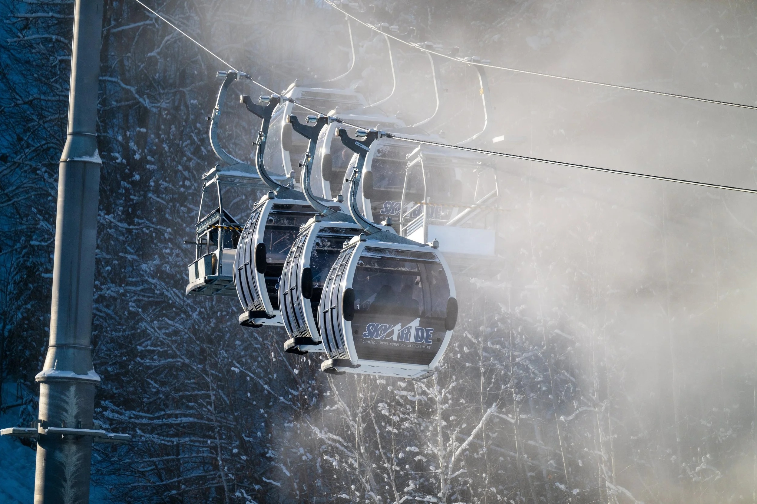 Ski gondola cabins suspended on cable above snow-covered forest and mountain slope in bright sunlight and mist.
