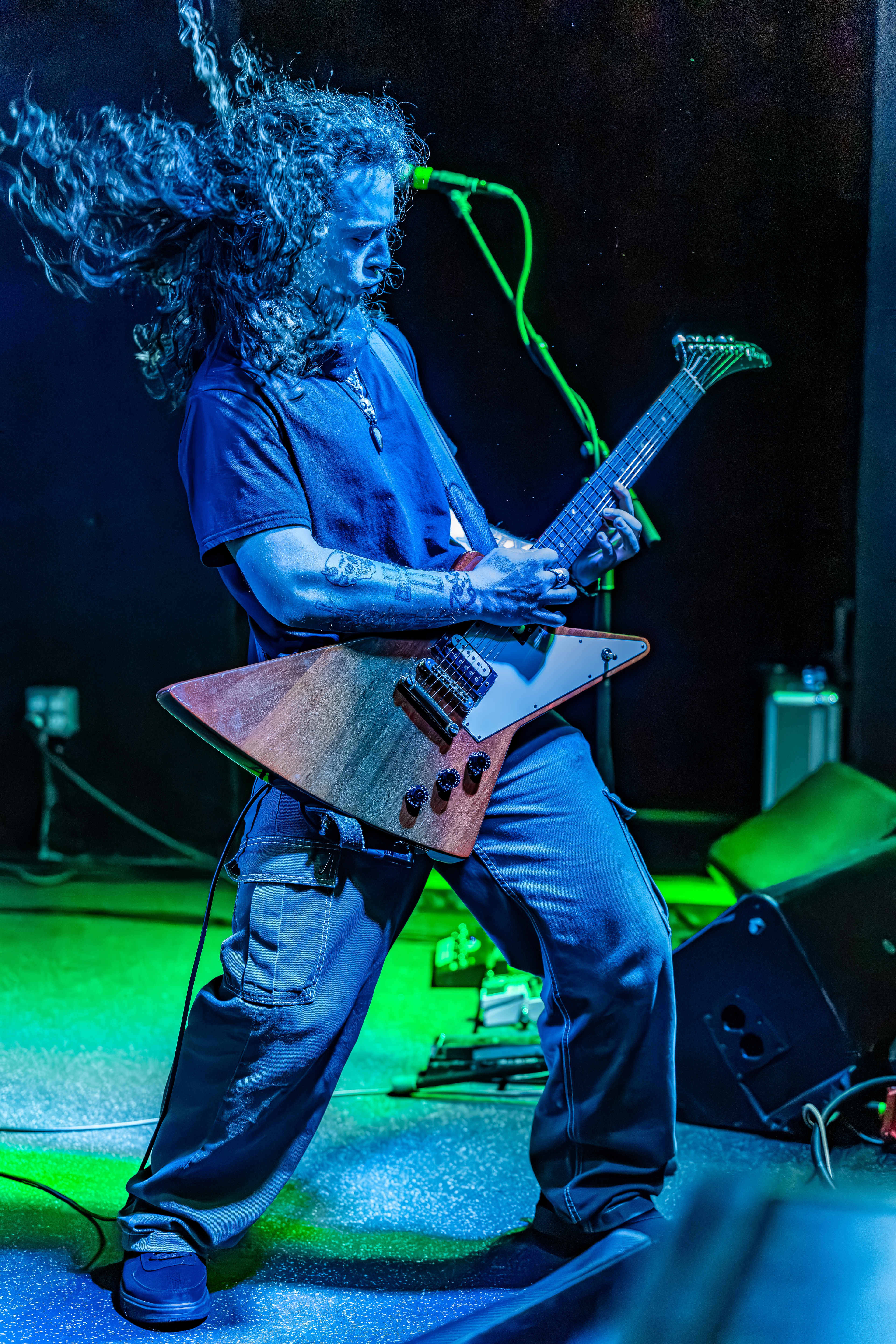 A guitarist with long curly hair plays an angular guitar on stage, bathed in blue and green stage lighting with a dark background.